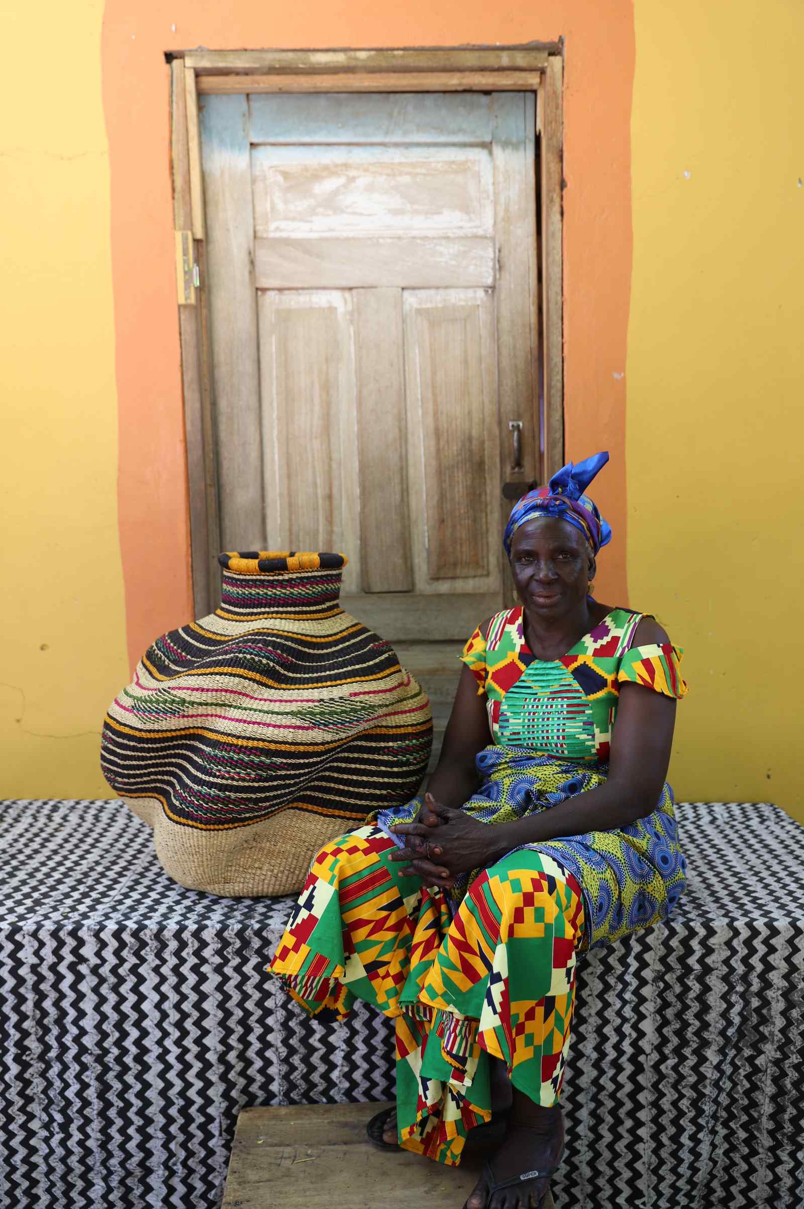 Medium handwoven Bolga art basket in natural straw with multicolour banded shoulder, beside a seated woman in kente.
