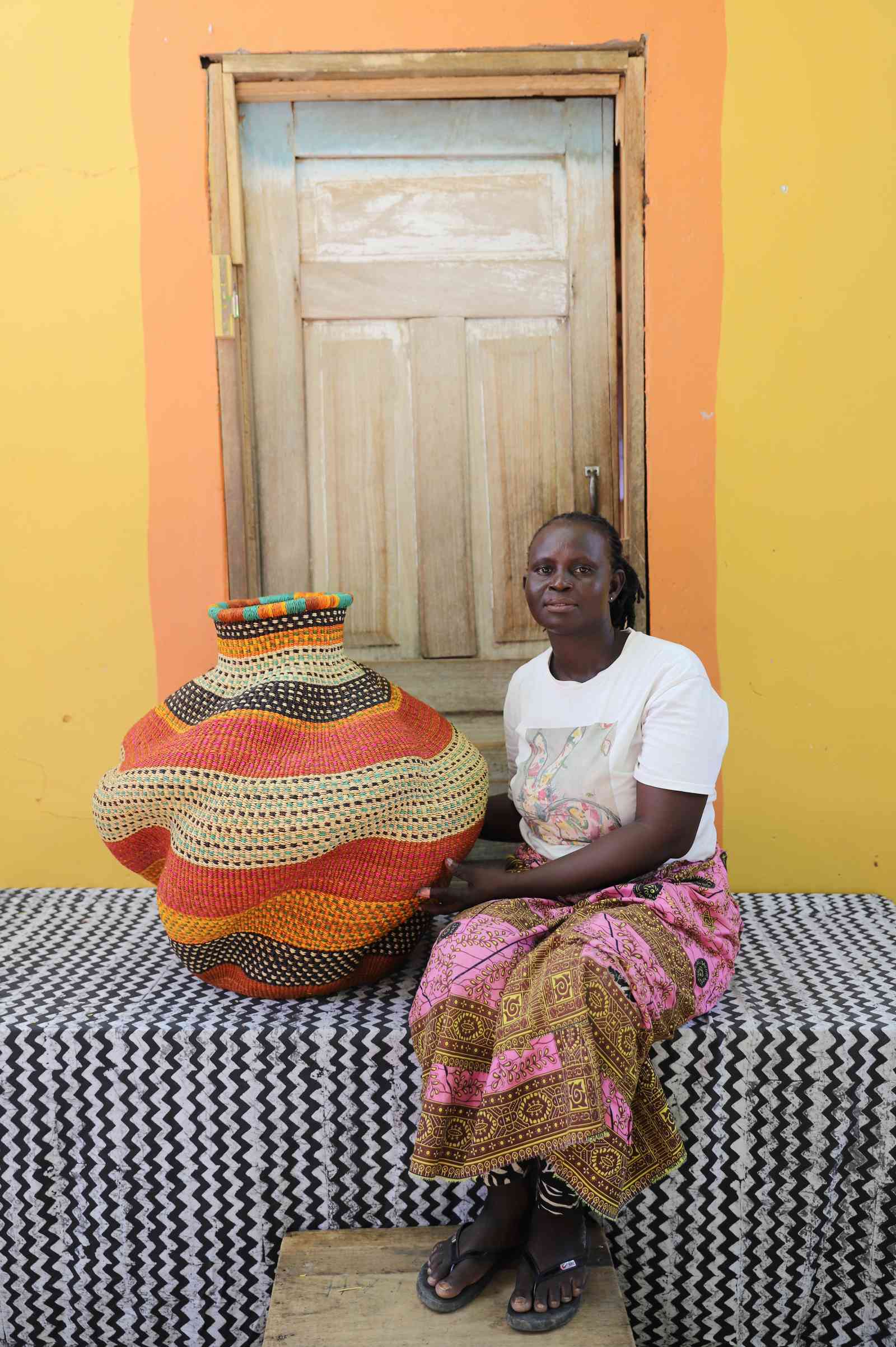 Medium handwoven Bolga art basket in rust-orange with dark dotted bands, beside a seated woman in white.