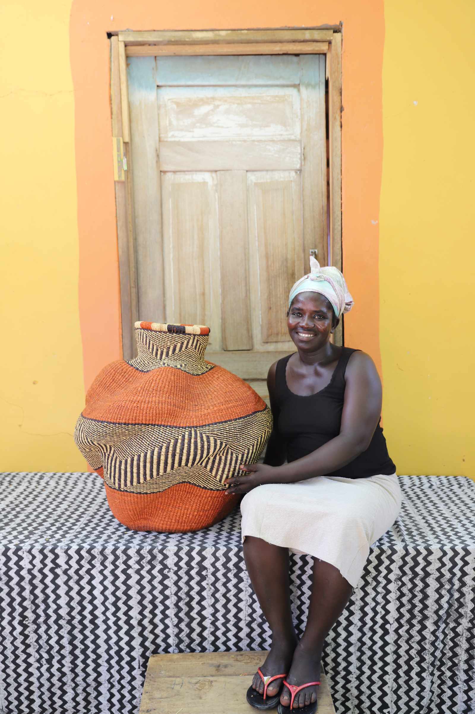 Medium handwoven Bolga art basket in rust-orange with dark spotted shoulder, beside a seated woman in black.