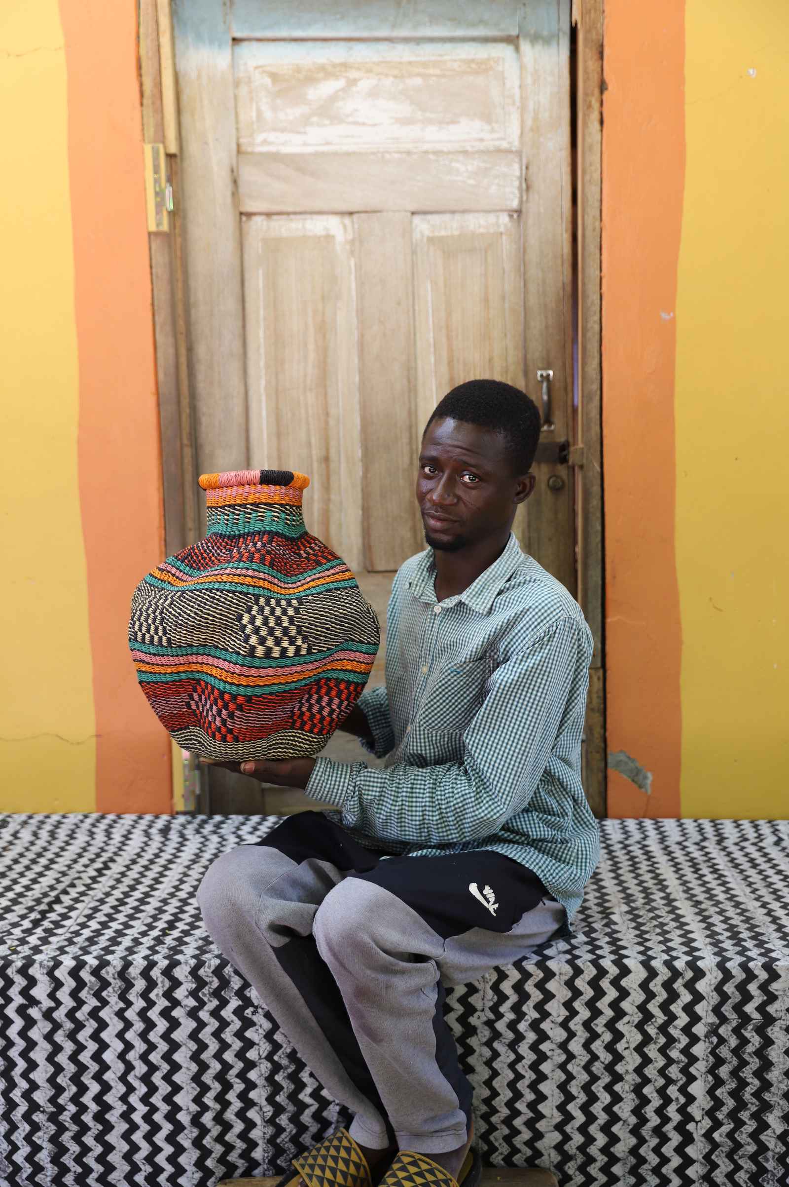 Handwoven Bolga art basket in natural straw with multicolour banded body, held by a seated man in checked shirt.