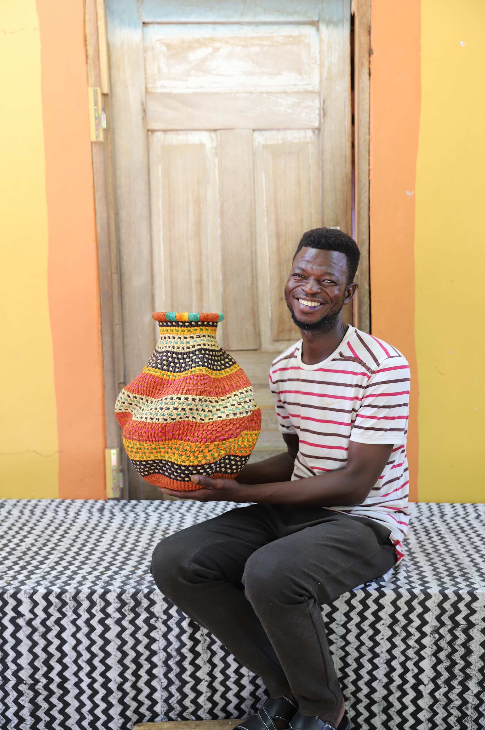 Handwoven Bolga art basket in rust and orange bands with dotted rim, held by a seated man in stripes.
