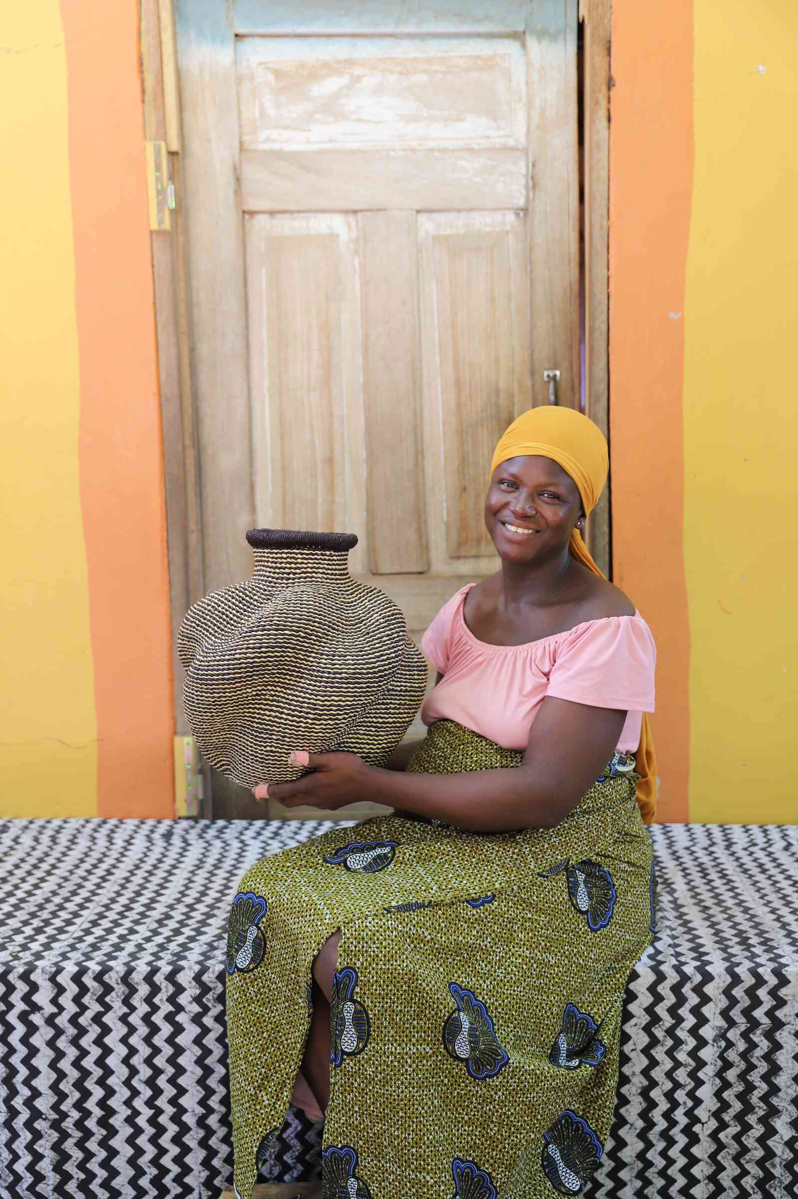 Handwoven Bolga art basket in natural straw with dark rim, held by a seated woman in pink and yellow.