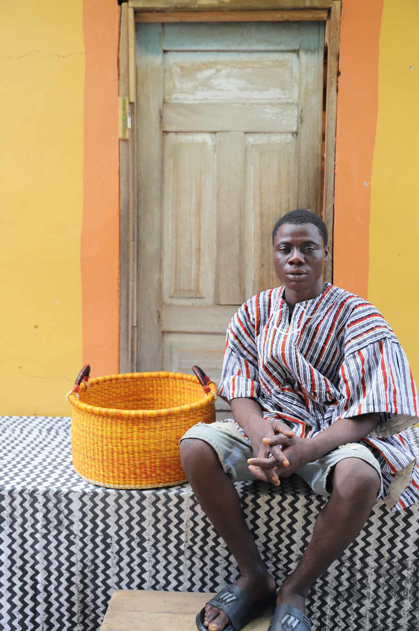 Small Bolga storage tub in natural with dark bands and orange centre, beside a seated woman in yellow.