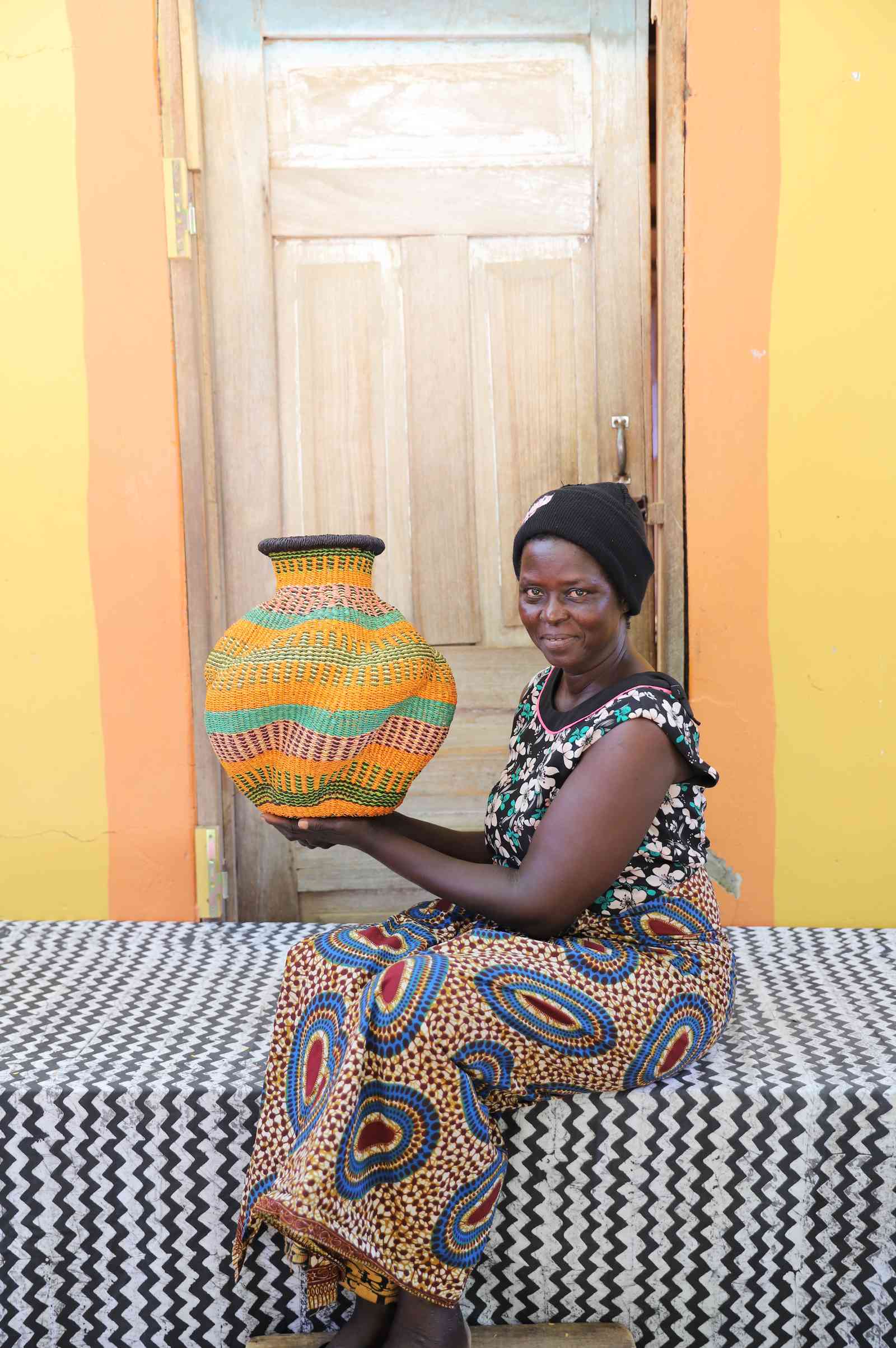 Handwoven Bolga art basket in natural straw with green, red and orange bands, held by a seated woman.