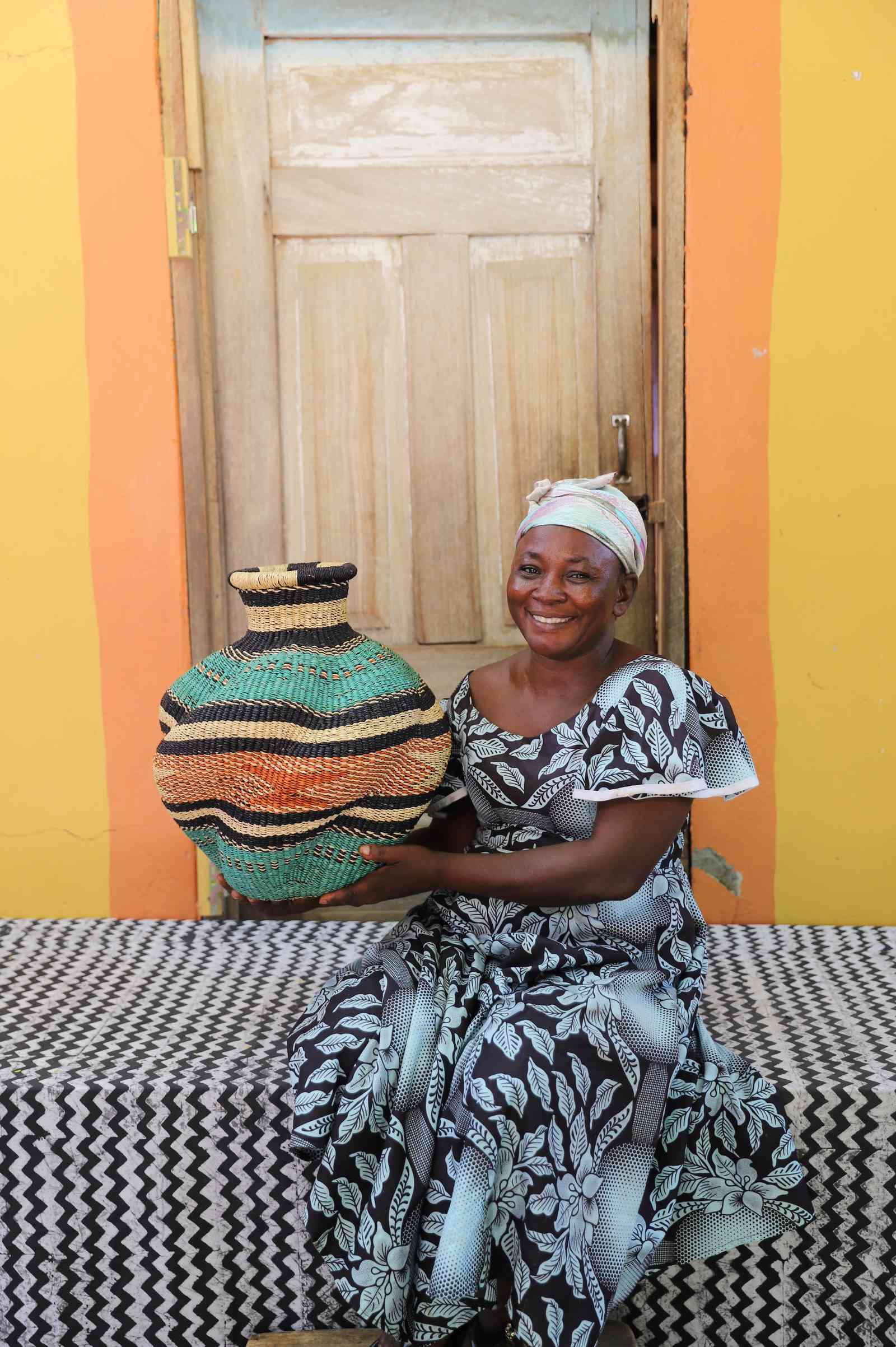 Handwoven Bolga art basket in teal with pink and orange banded shoulder, held by a seated woman in patterned dress.