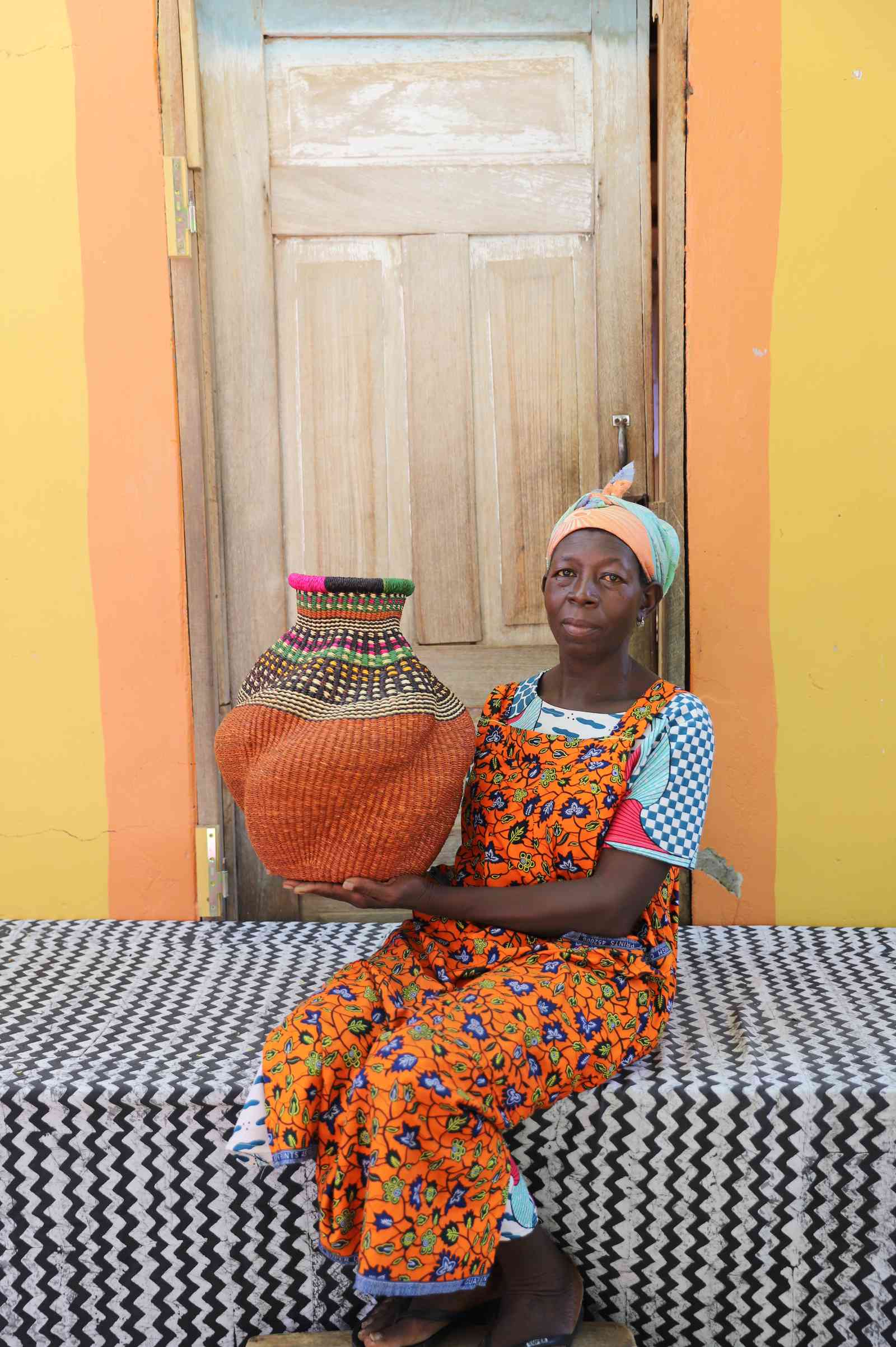 Handwoven Bolga art basket in rust-orange with multicolour banded shoulder, held by a seated woman in floral.