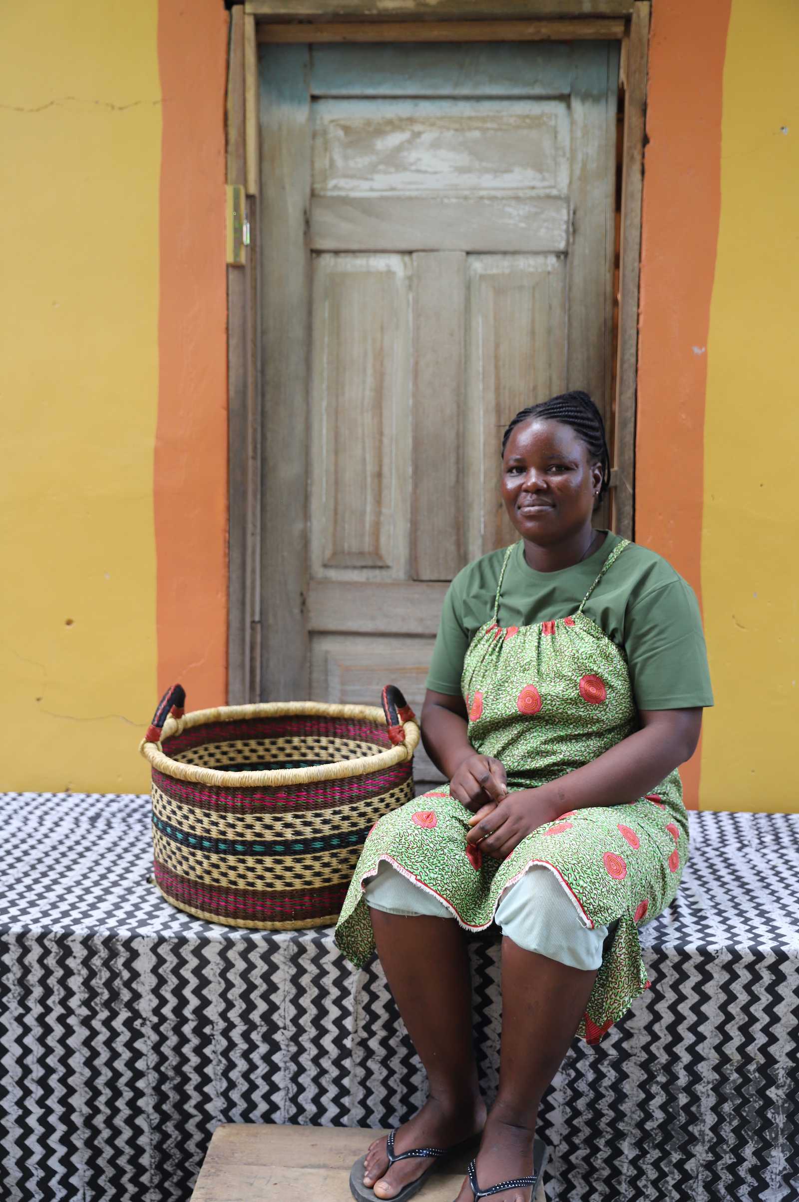 Small Bolga storage tub in natural with red and dark bands, beside a seated woman in a printed dress.