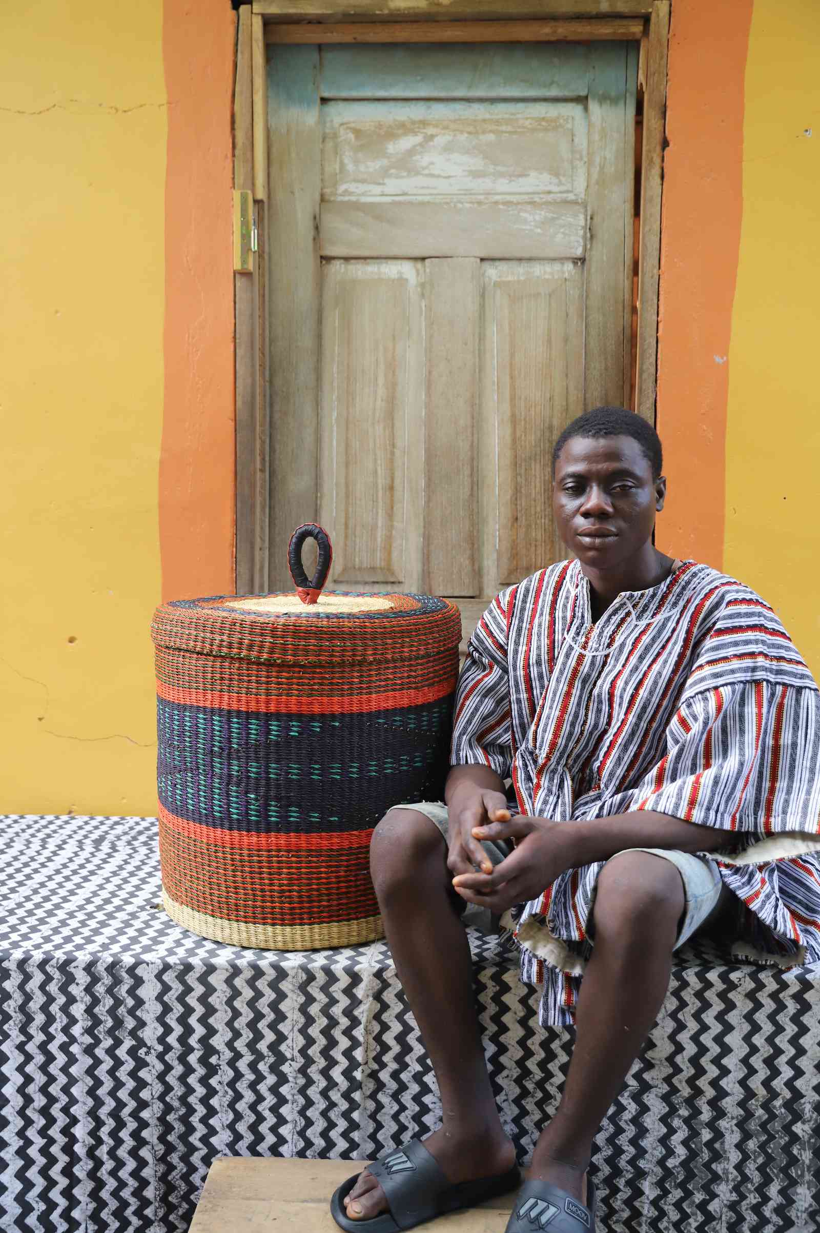 Large Bolga laundry basket in rust-orange with dark band, beside a seated man in a striped tunic.