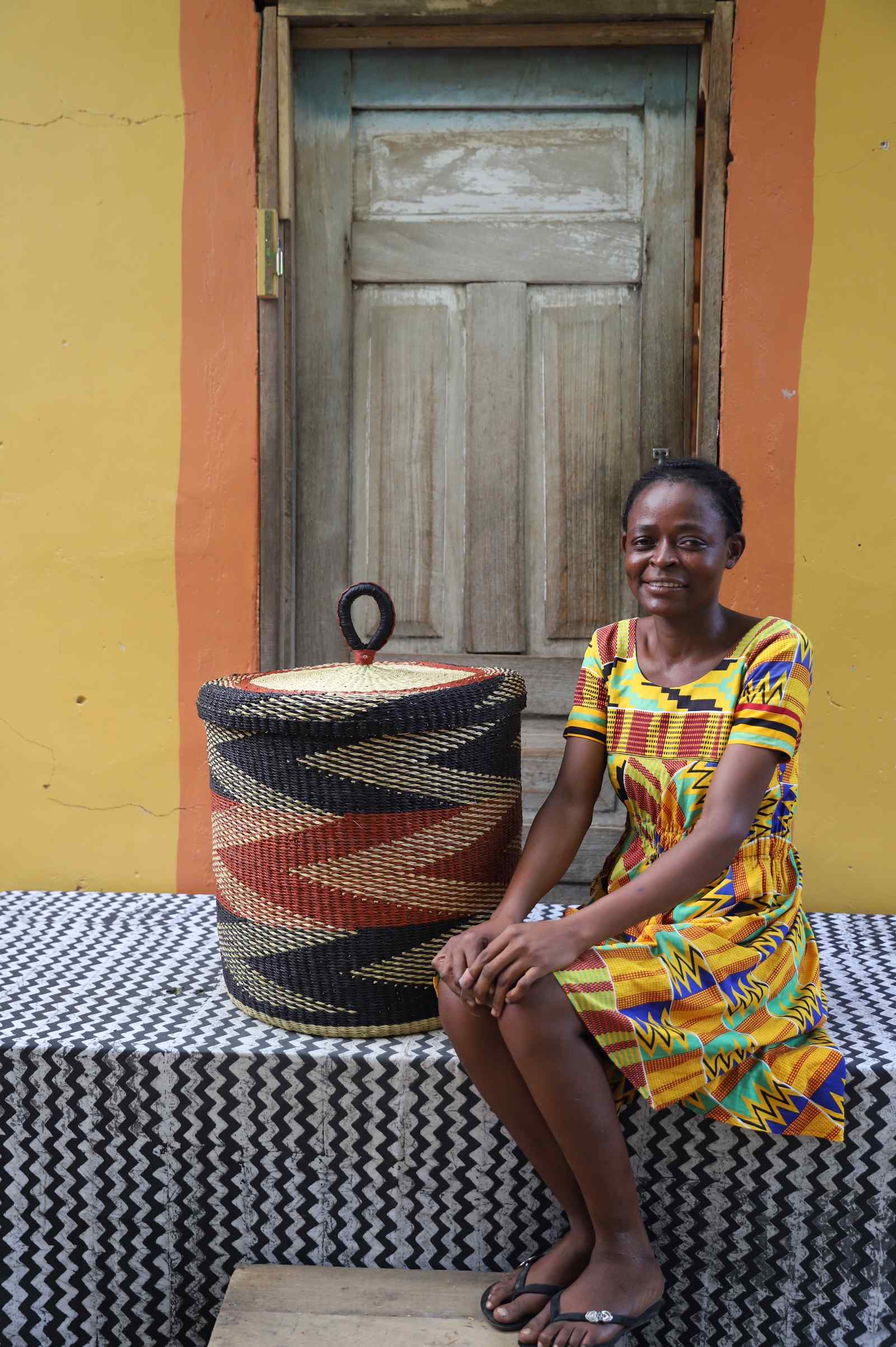 Medium Bolga laundry basket in rust-red and black bands, beside a seated woman in a printed dress.