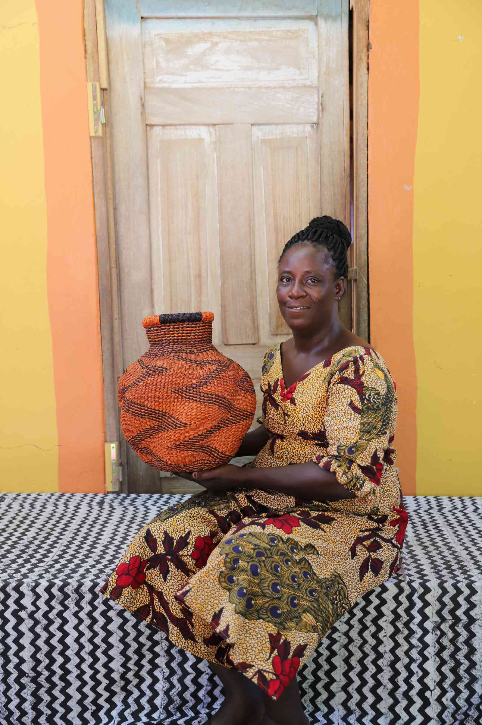 Handwoven Bolga art basket in rust-orange with dark zigzag wave motif, held by a seated woman in a printed dress.