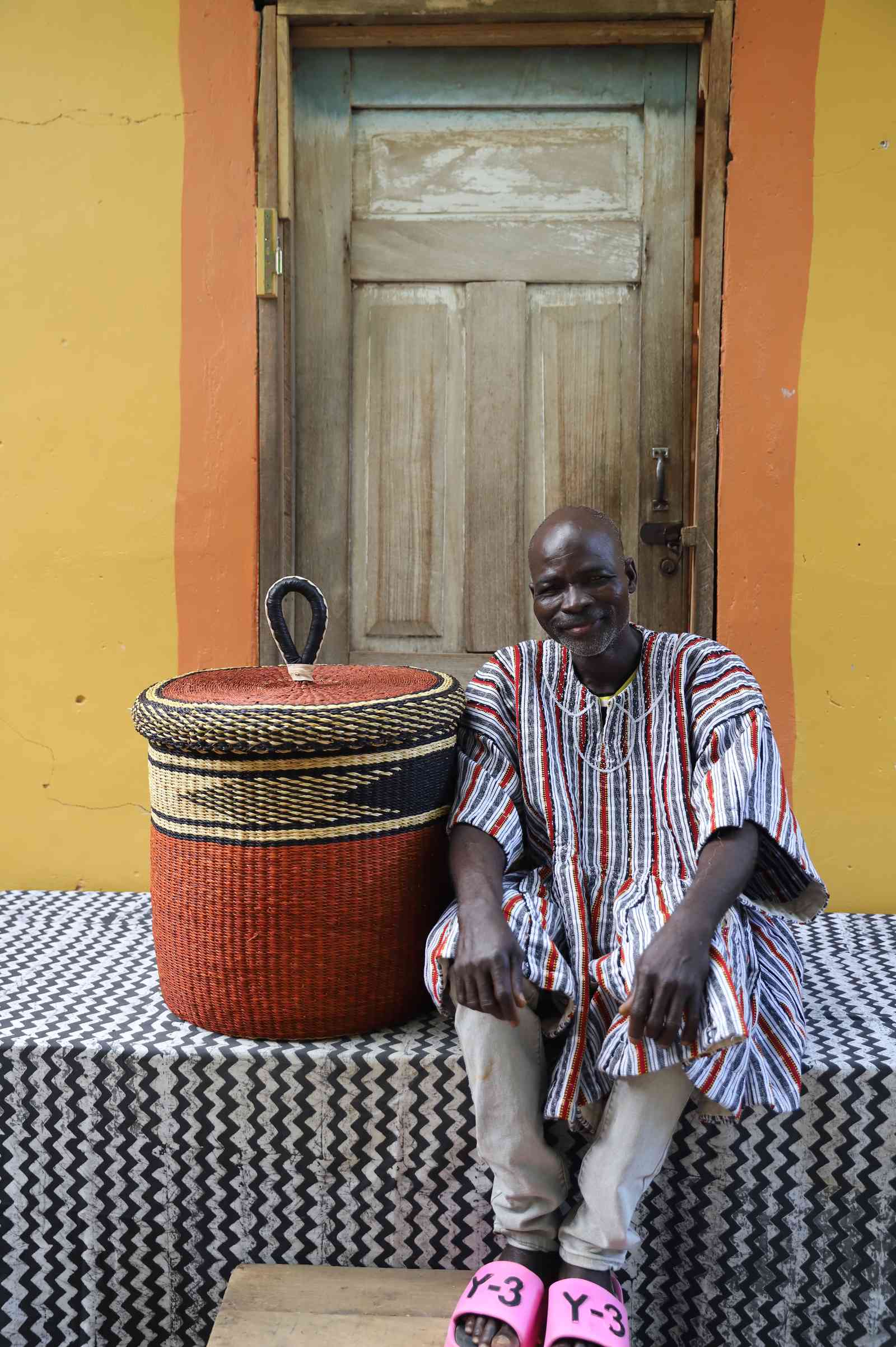 Medium Bolga laundry basket with dark top and rust-orange base, beside a seated man in a striped tunic.