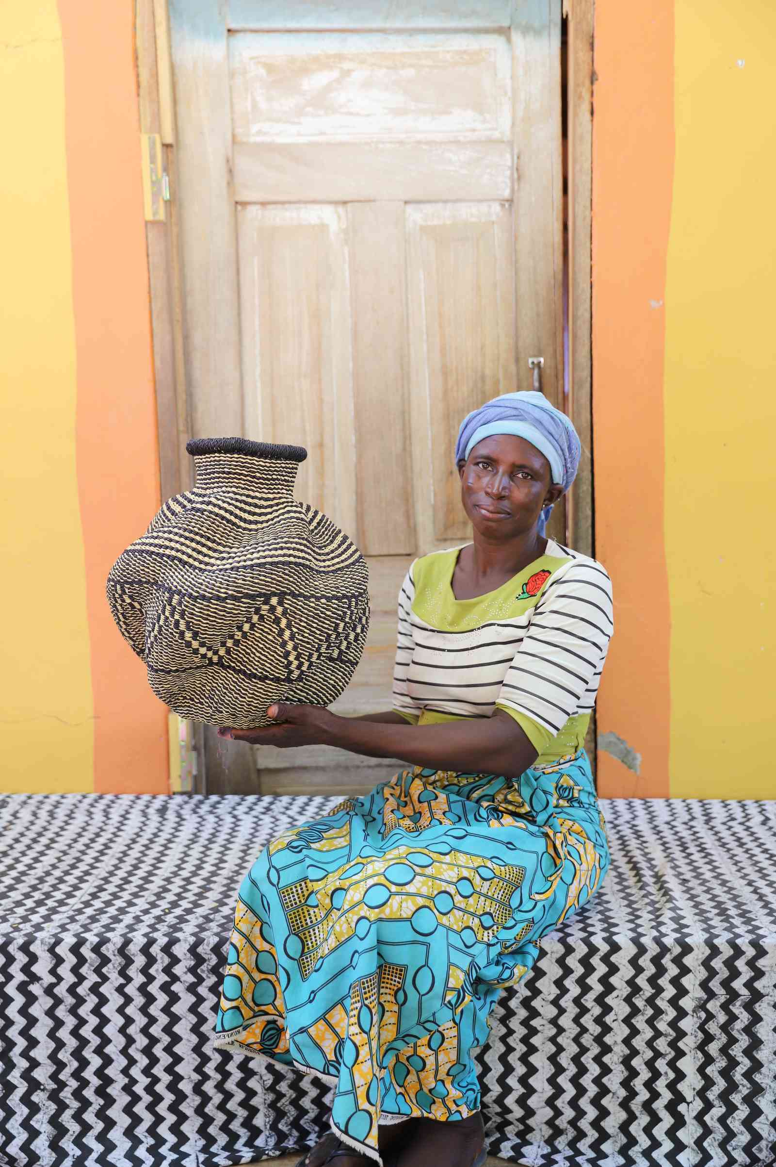 Handwoven Bolga art basket in natural straw with fine dark diamond weave, held by a seated woman.