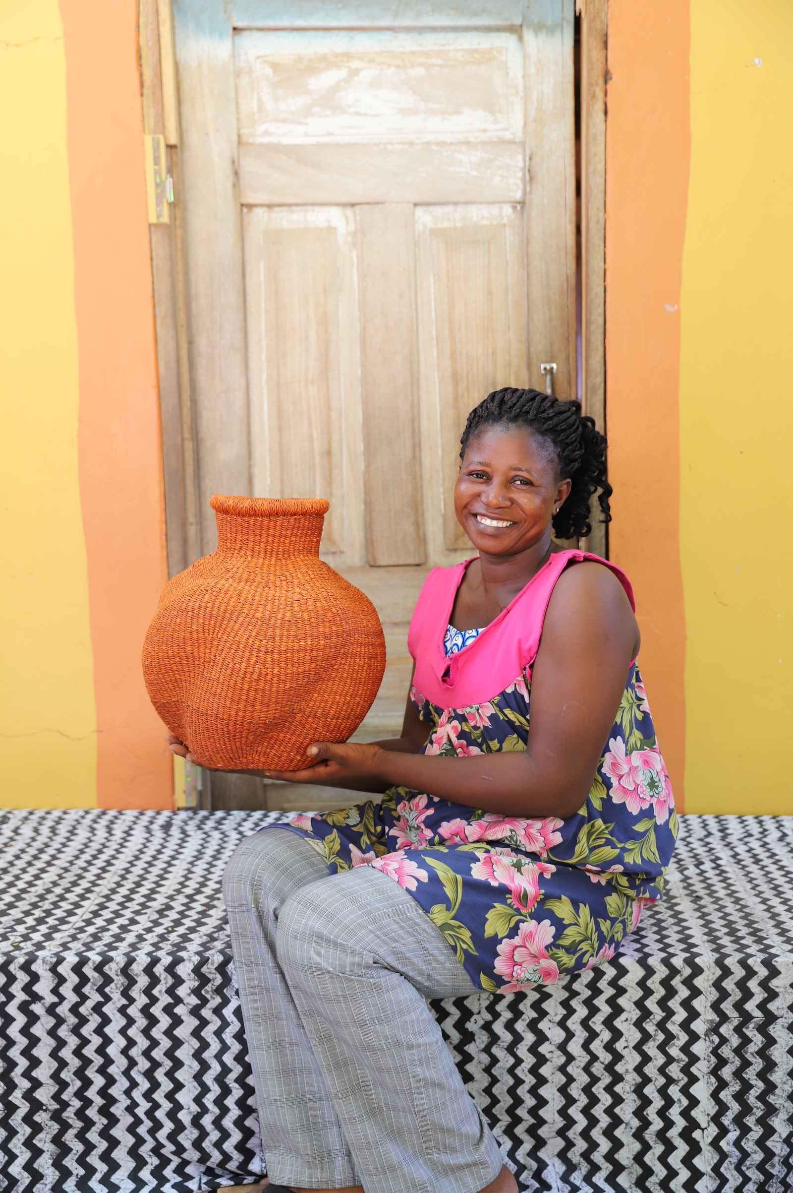 Handwoven Bolga art basket in dyed rust-orange with a dark rim, held by a seated woman in pink and grey.