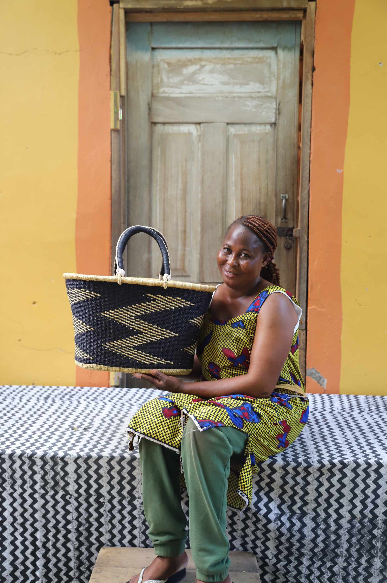 Large Bolga Zaane Tia market basket in black with natural zigzag, held by a seated woman in a patterned dress.