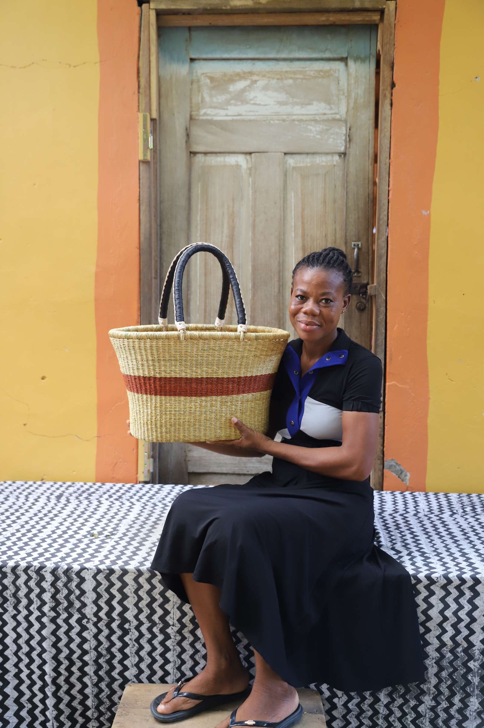 Large Bolga Zaane Tia market basket with rust-orange band, held by a seated woman in a black dress.