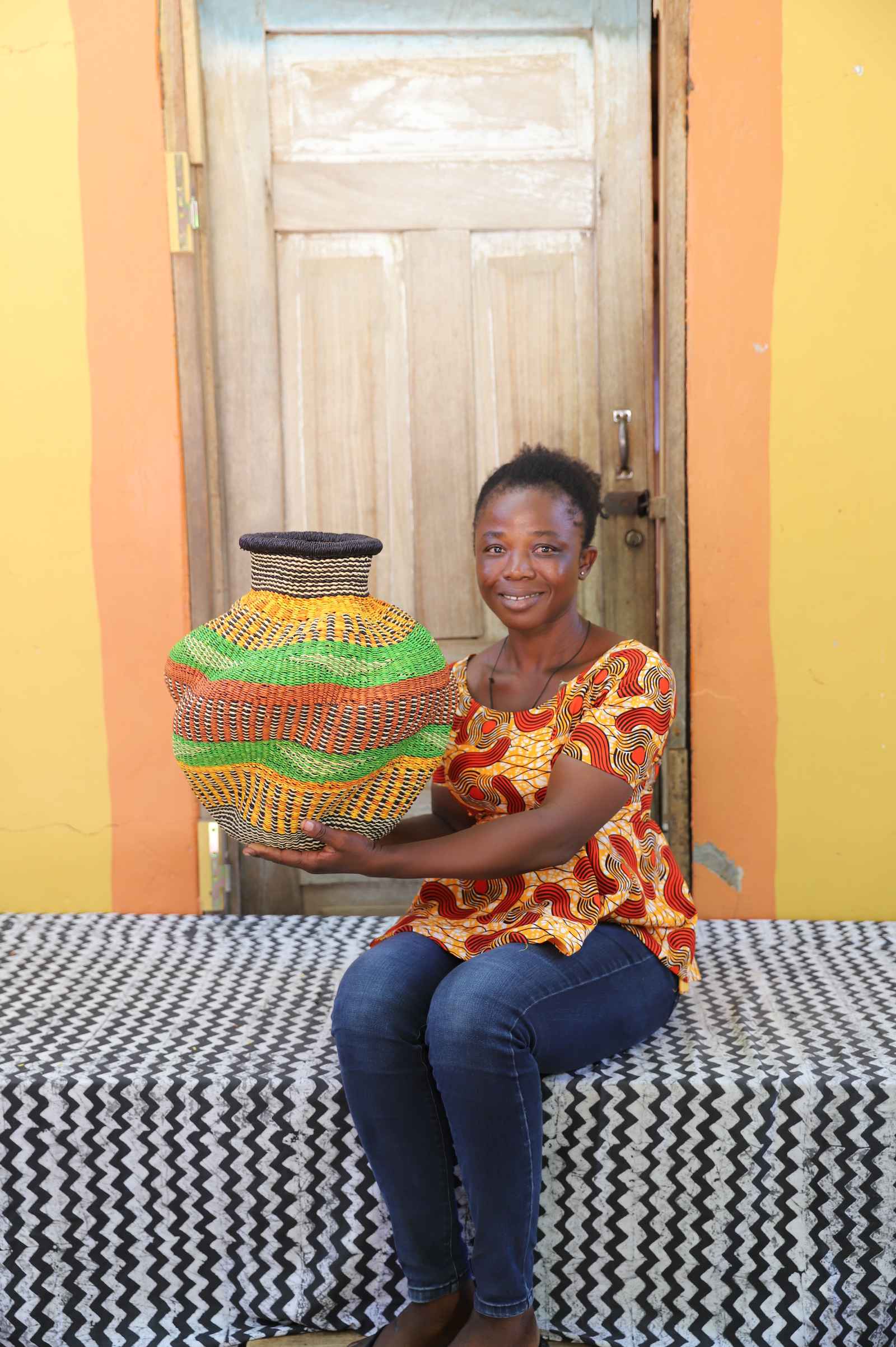 Handwoven Bolga art basket in green, orange and yellow bands, held by a seated woman in an orange top.