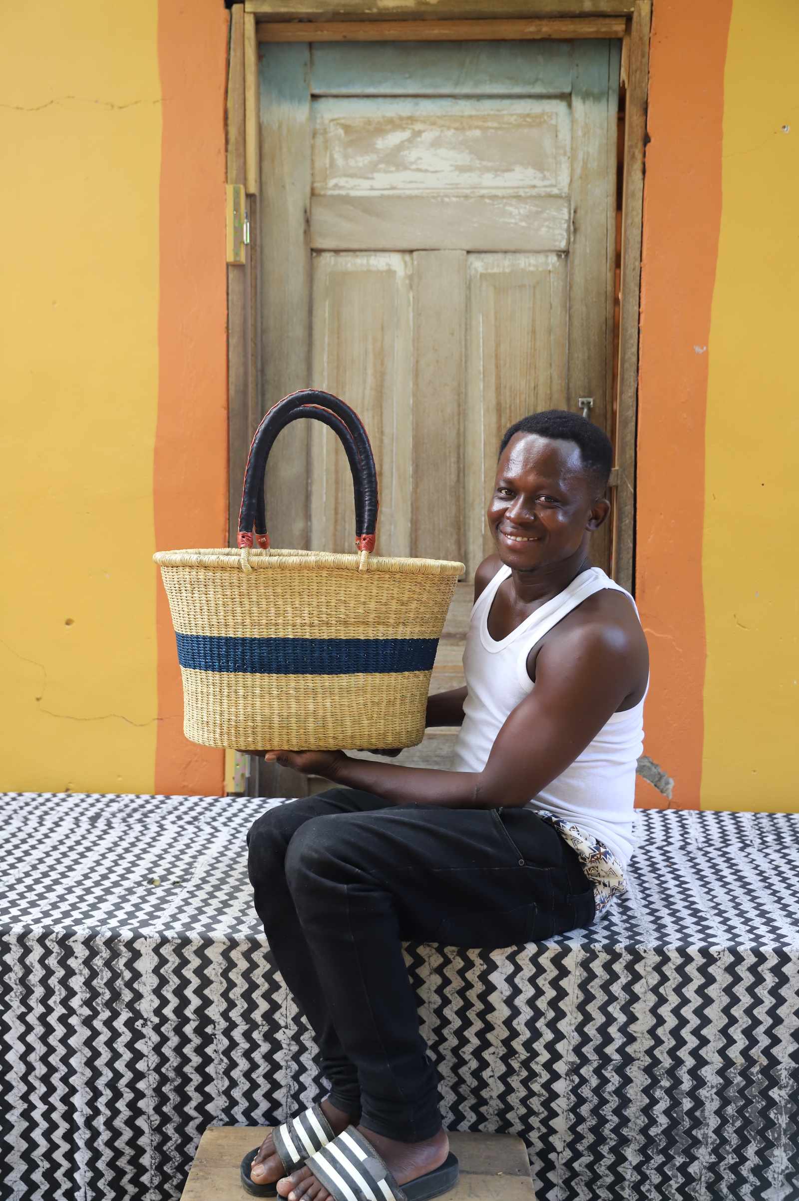 Large Bolga Zaane Tia market basket with navy band, held by a seated man in a white vest.