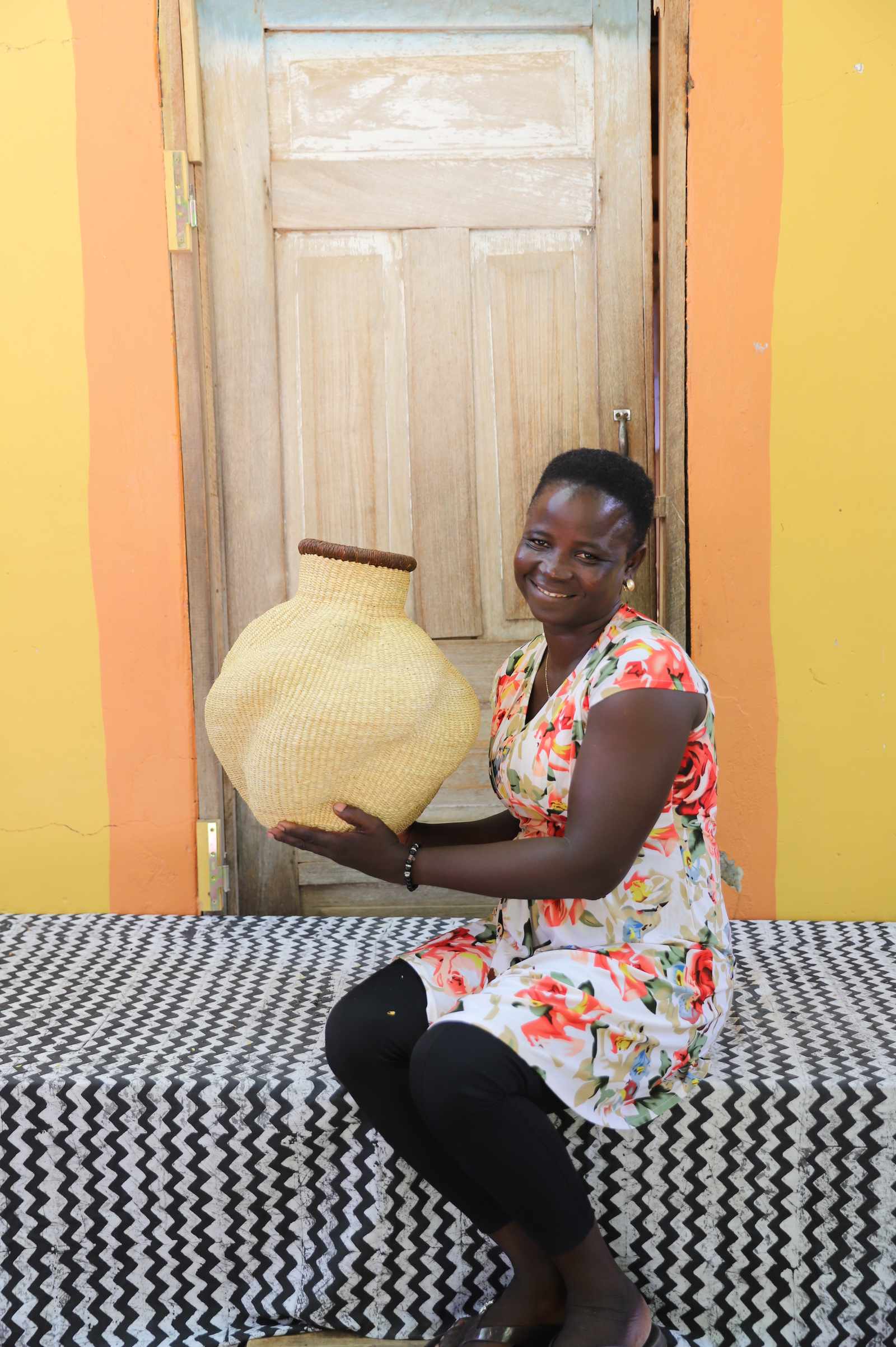 Handwoven Bolga art basket in natural straw with a dark rim, held by a seated woman in a floral dress.