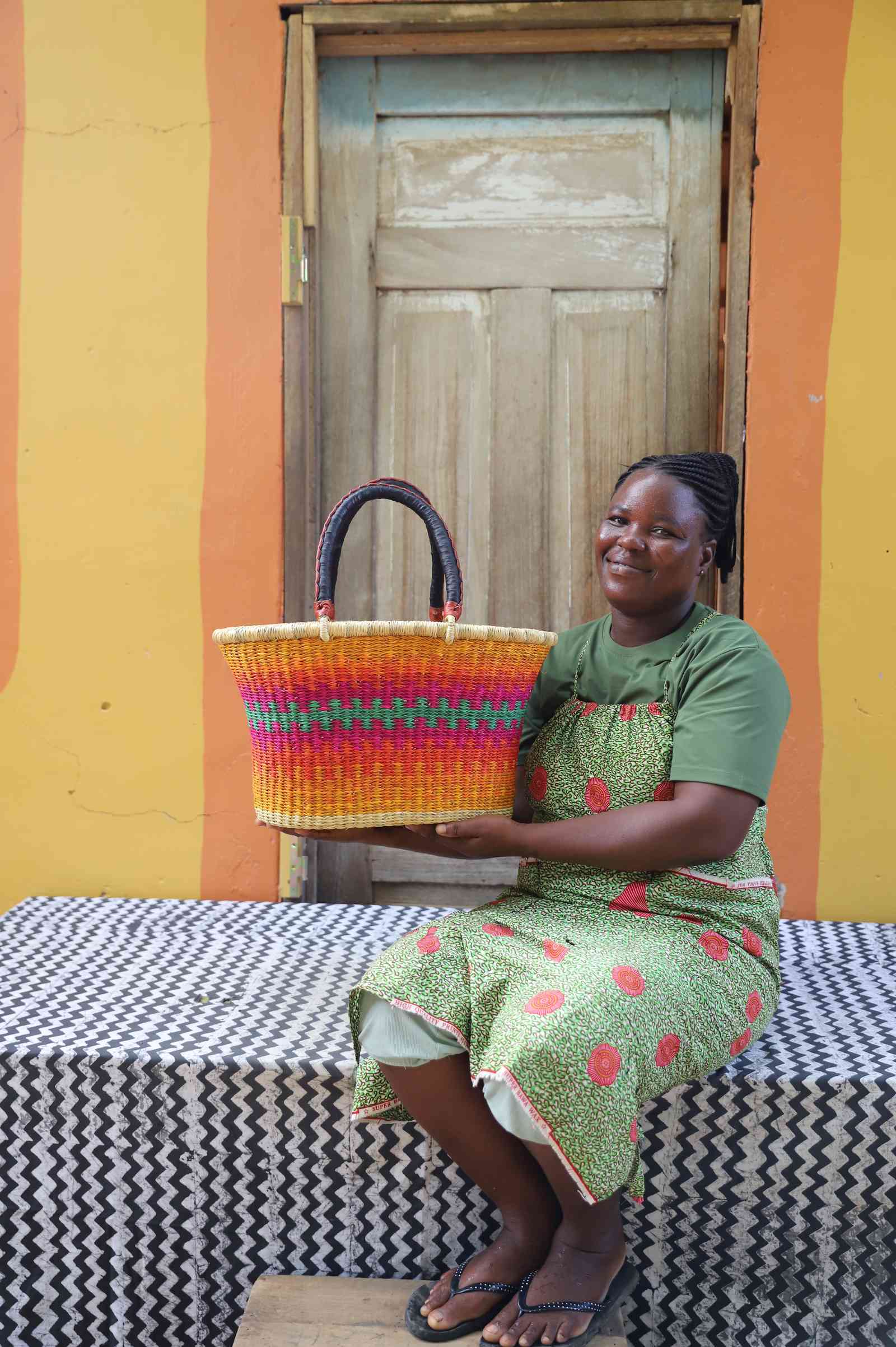 Large Bolga Zaane Tia market basket in multicolour stripes, held by a seated woman in green.