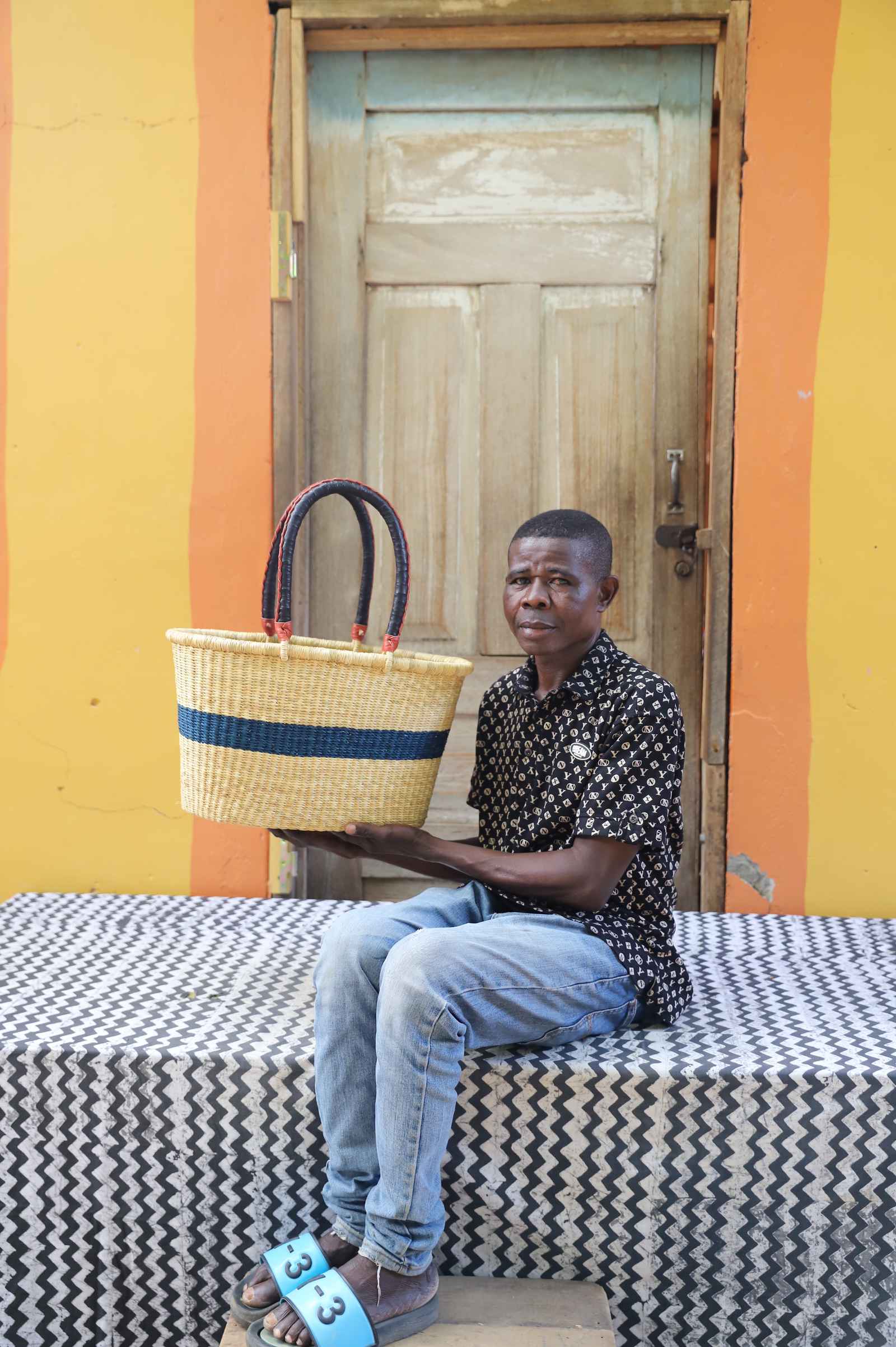 Medium Bolga Zaane Tia market basket with slim navy band, held by a seated man in a dark top.