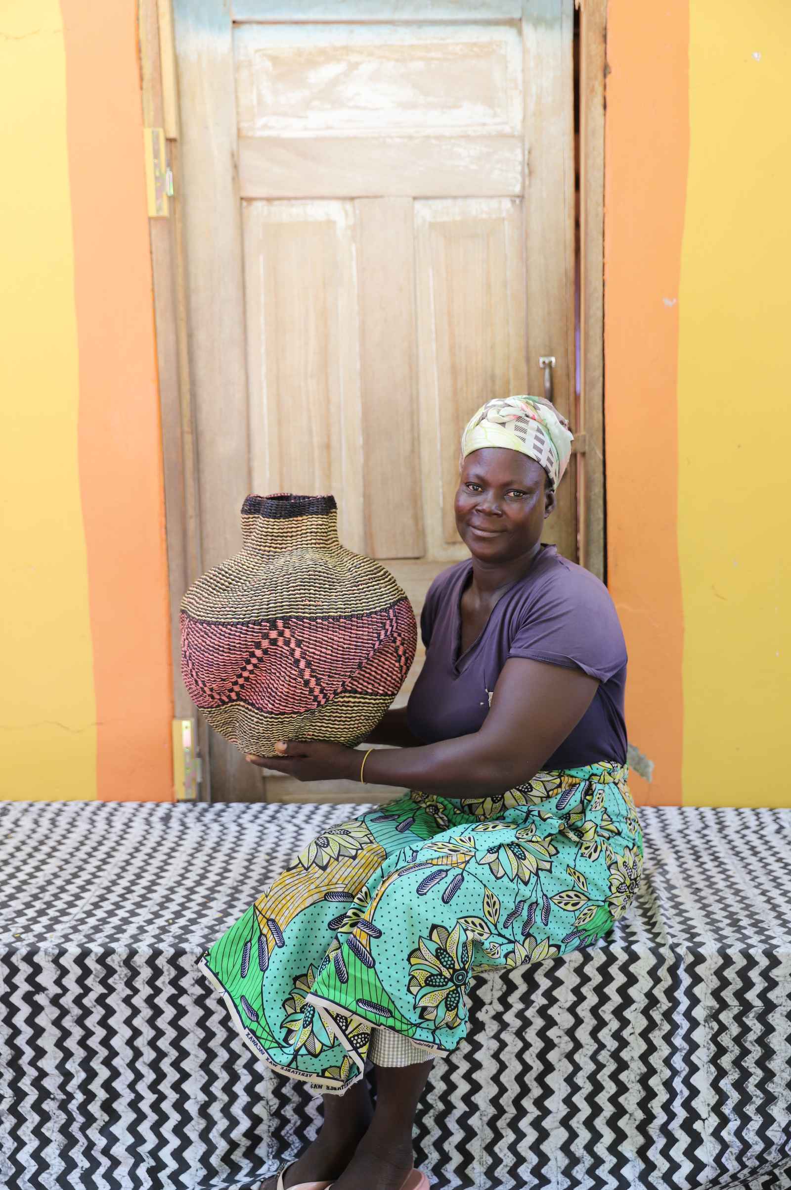 Handwoven Bolga art basket in natural straw with dark diamond weave and red centre, held by a seated woman.