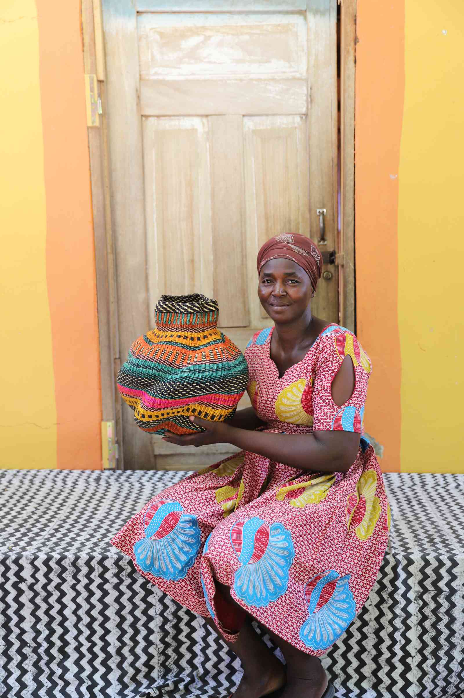 Handwoven Bolga art basket with fine multicolour stripes and natural rim, held by a seated woman in red and blue.