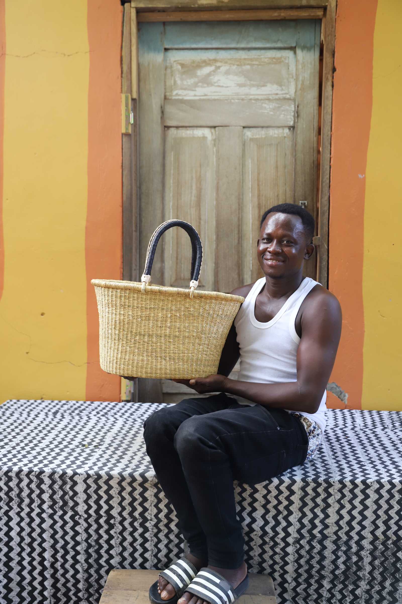 Medium Bolga Zaane Tia market basket in plain natural straw, held by a seated man in white.