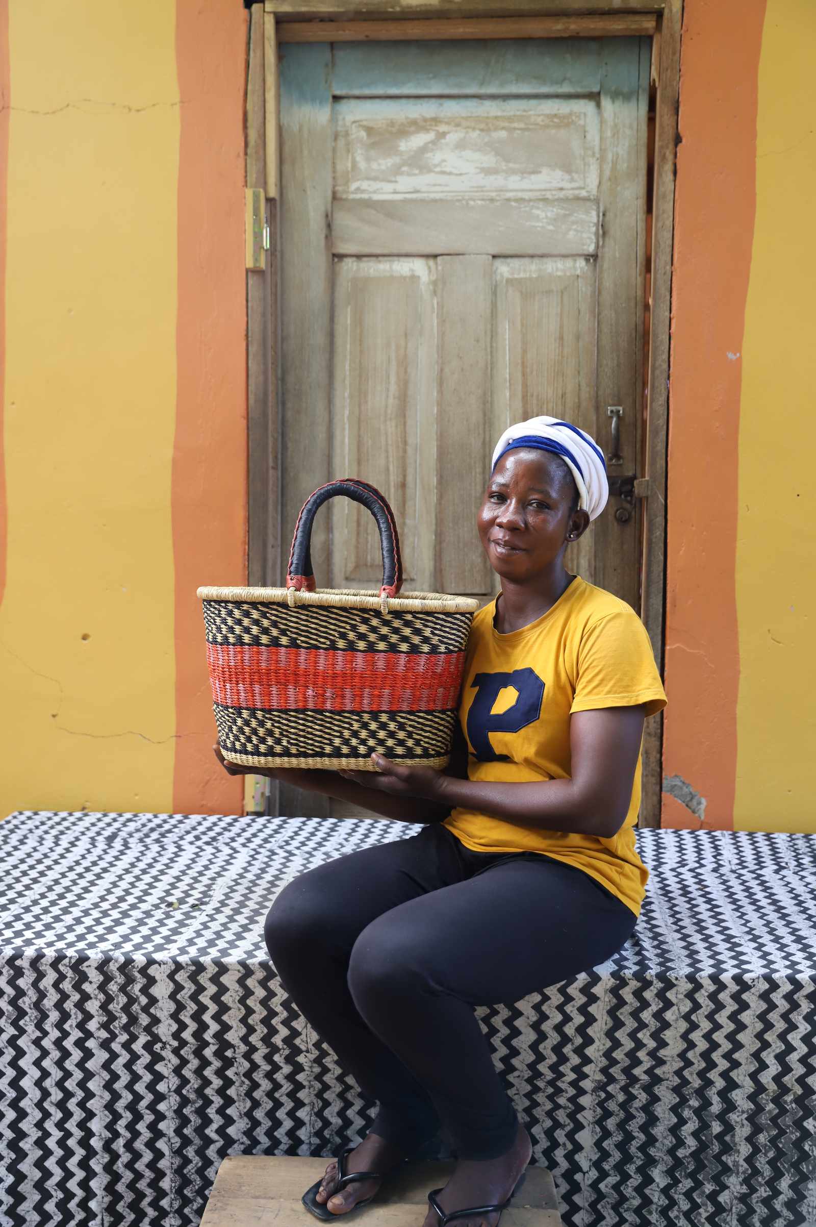 Medium Bolga Zaane Tia market basket with red and orange bands, held by a seated woman in yellow.