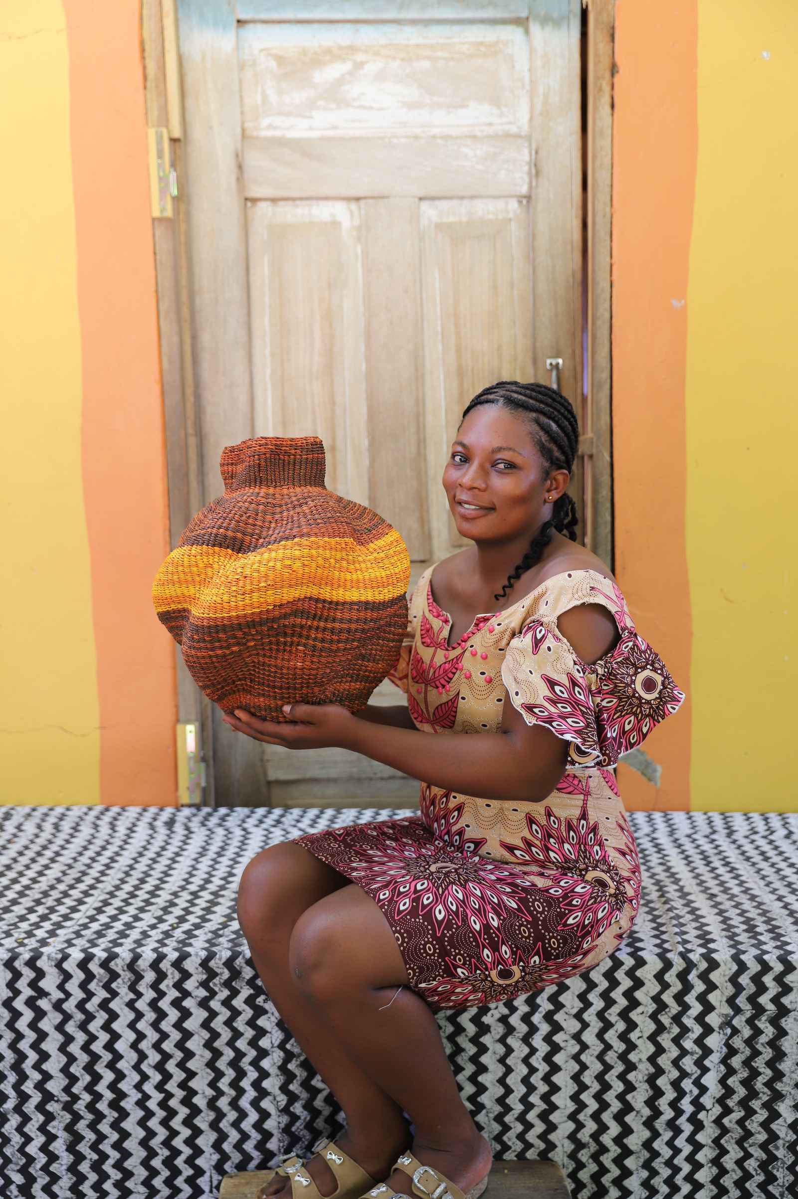 Handwoven Bolga art basket in rust-red with a yellow and orange band, held by a seated woman in pink.
