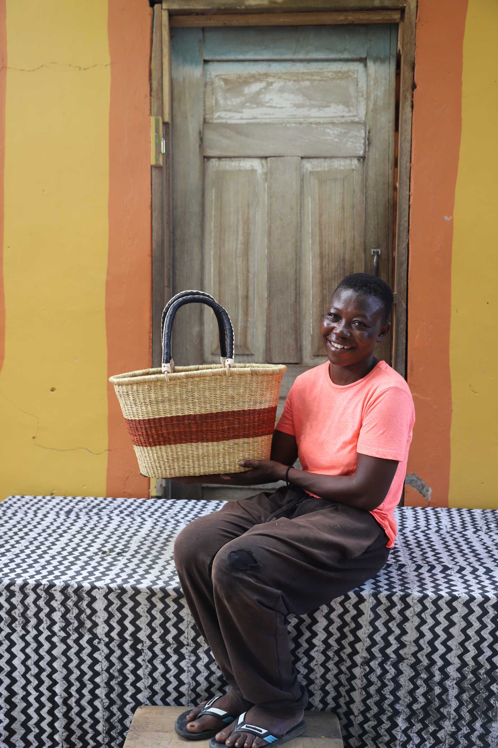 Medium Bolga Zaane Tia market basket with rust-orange band, held by a seated woman in coral.