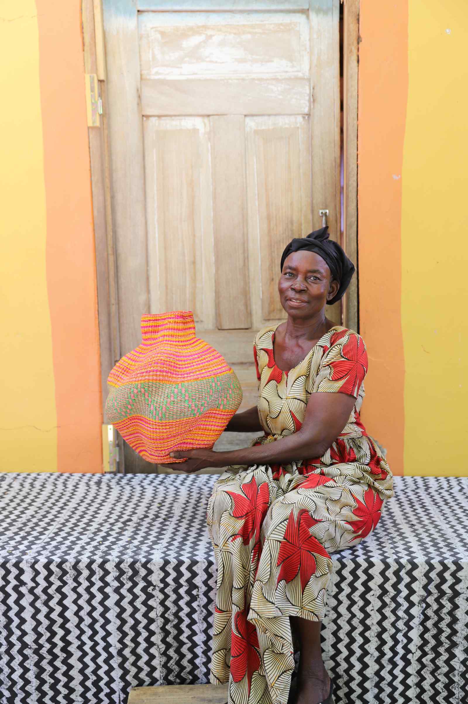 Handwoven Bolga art basket with fine pink and orange bands, held by a seated woman in red and cream.