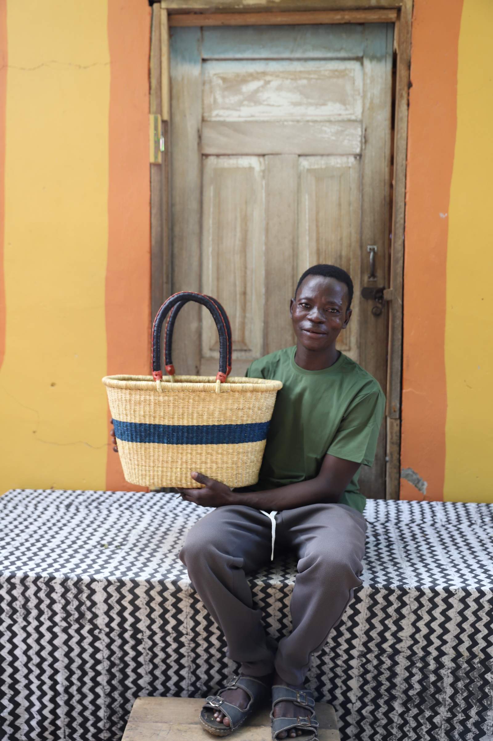 Medium Bolga Zaane Tia market basket with navy band, held by a seated man in a printed shirt.