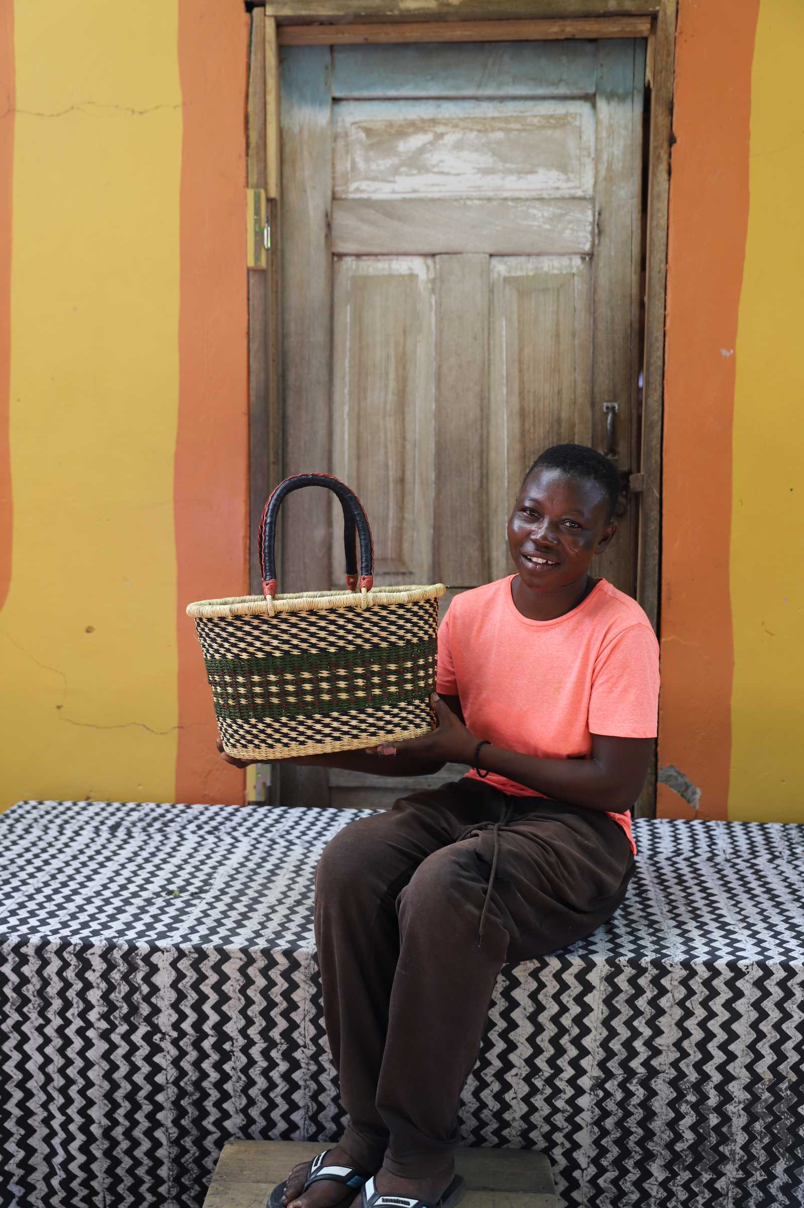 Small Bolga Zaane Tia market basket with dark and orange zigzag band, held by a seated woman in yellow.