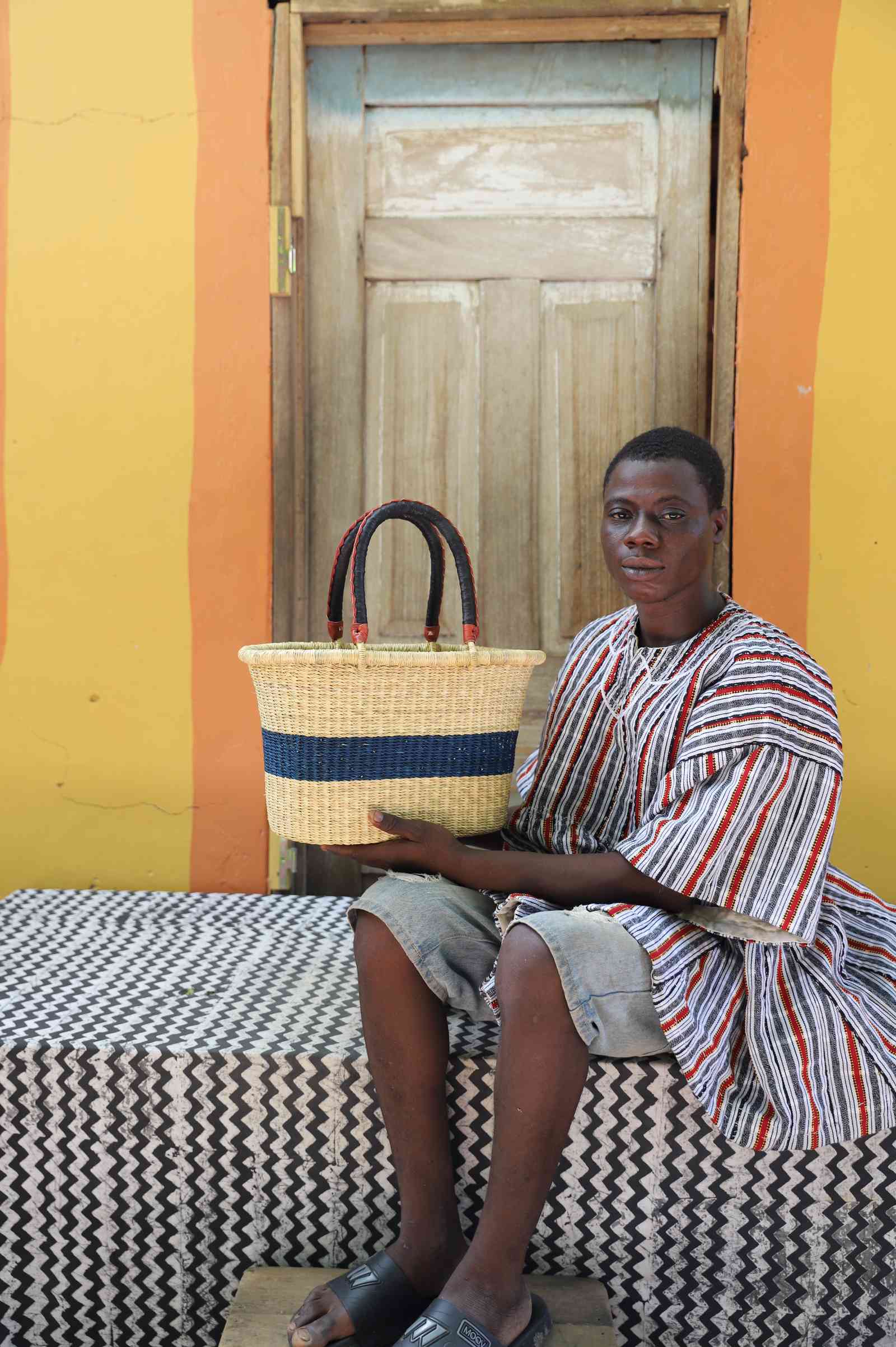 Small Bolga Zaane Tia market basket with navy band, held by a seated man in a striped tunic.