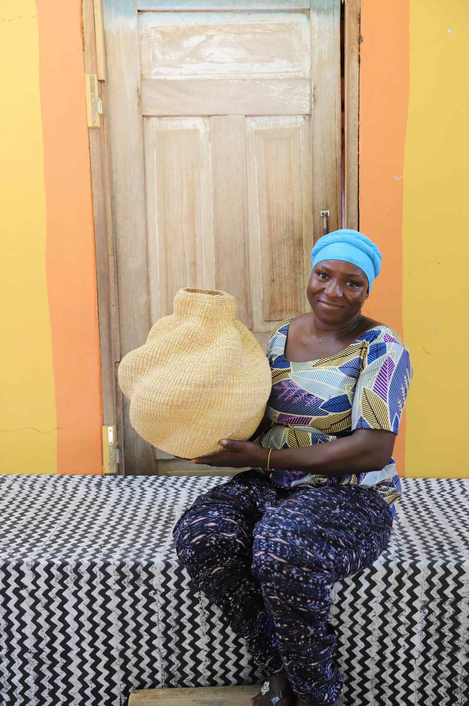 Handwoven Bolga art basket in an asymmetric natural straw form, held by a seated woman in blue and yellow.