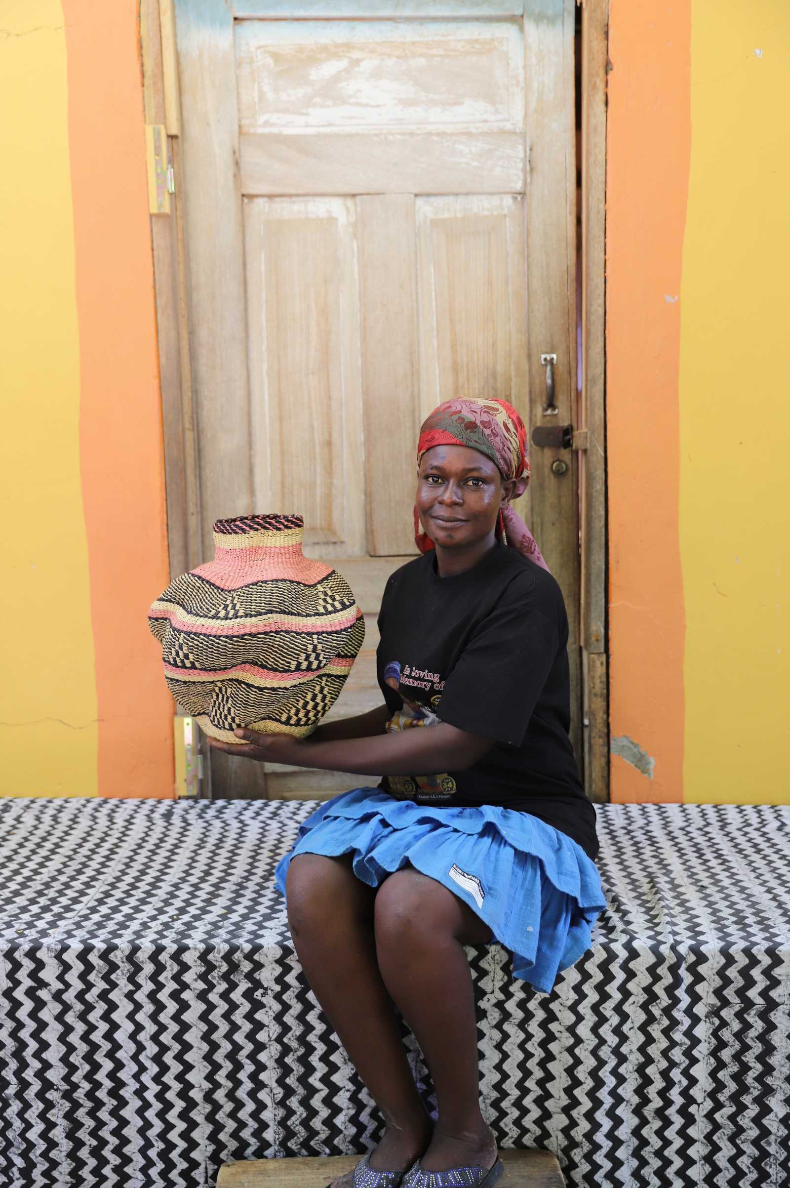 Handwoven Bolga art basket in natural straw with pink and dark bands, held by a seated woman in a red headscarf.