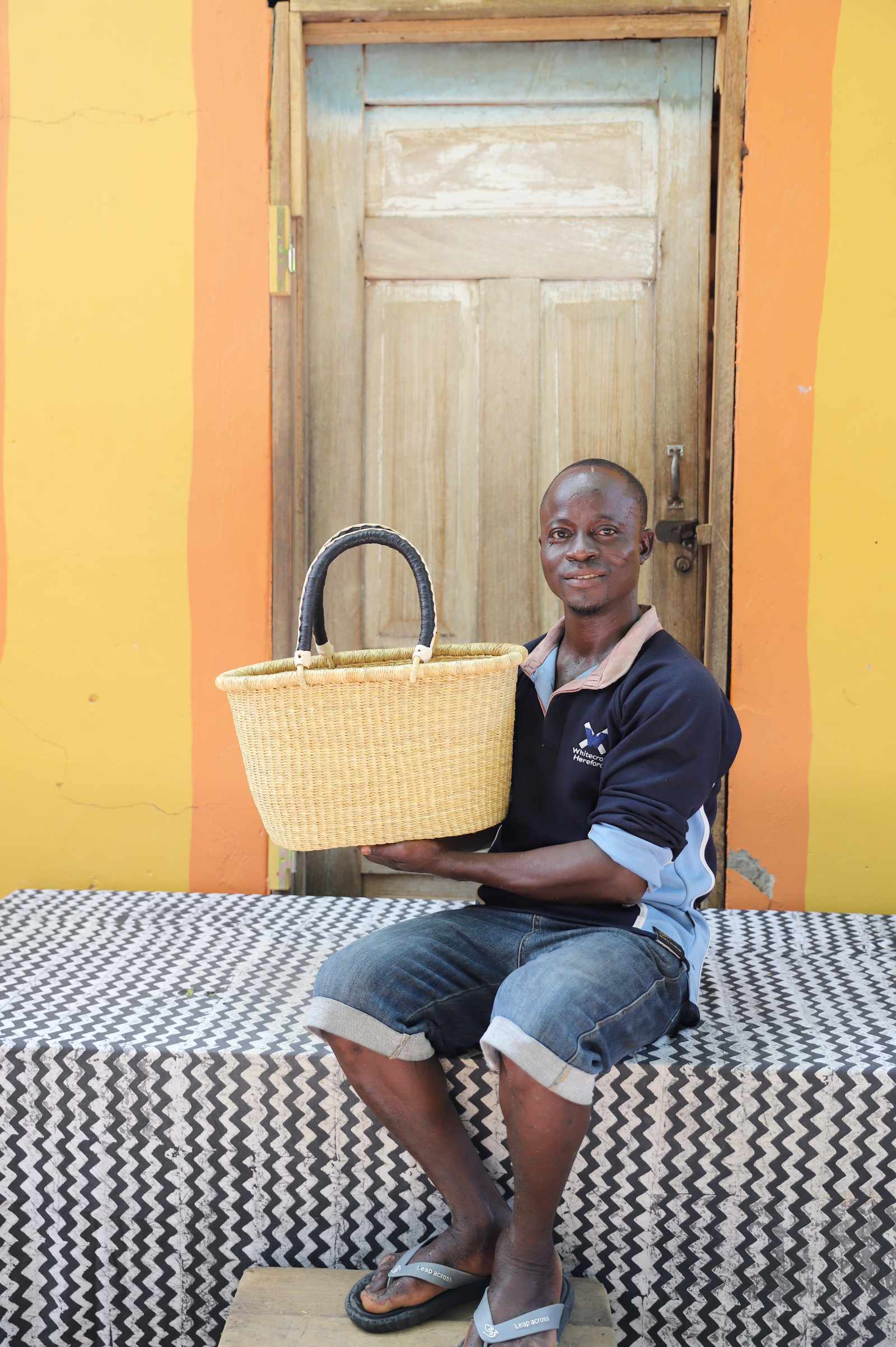 Small Bolga Zaane Tia market basket in plain natural straw, held by a seated man in a dark top.