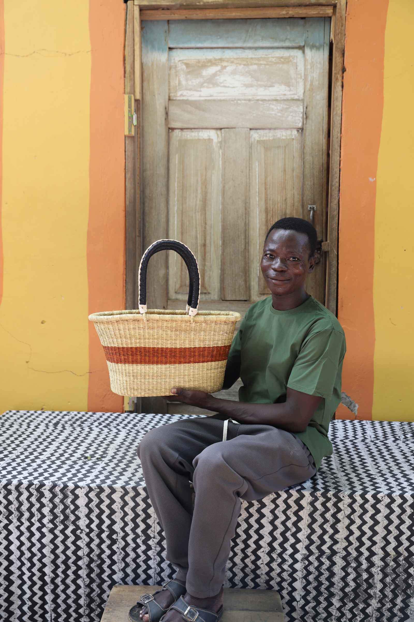Small Bolga Zaane Tia market basket with rust-orange band, held by a seated man in green.