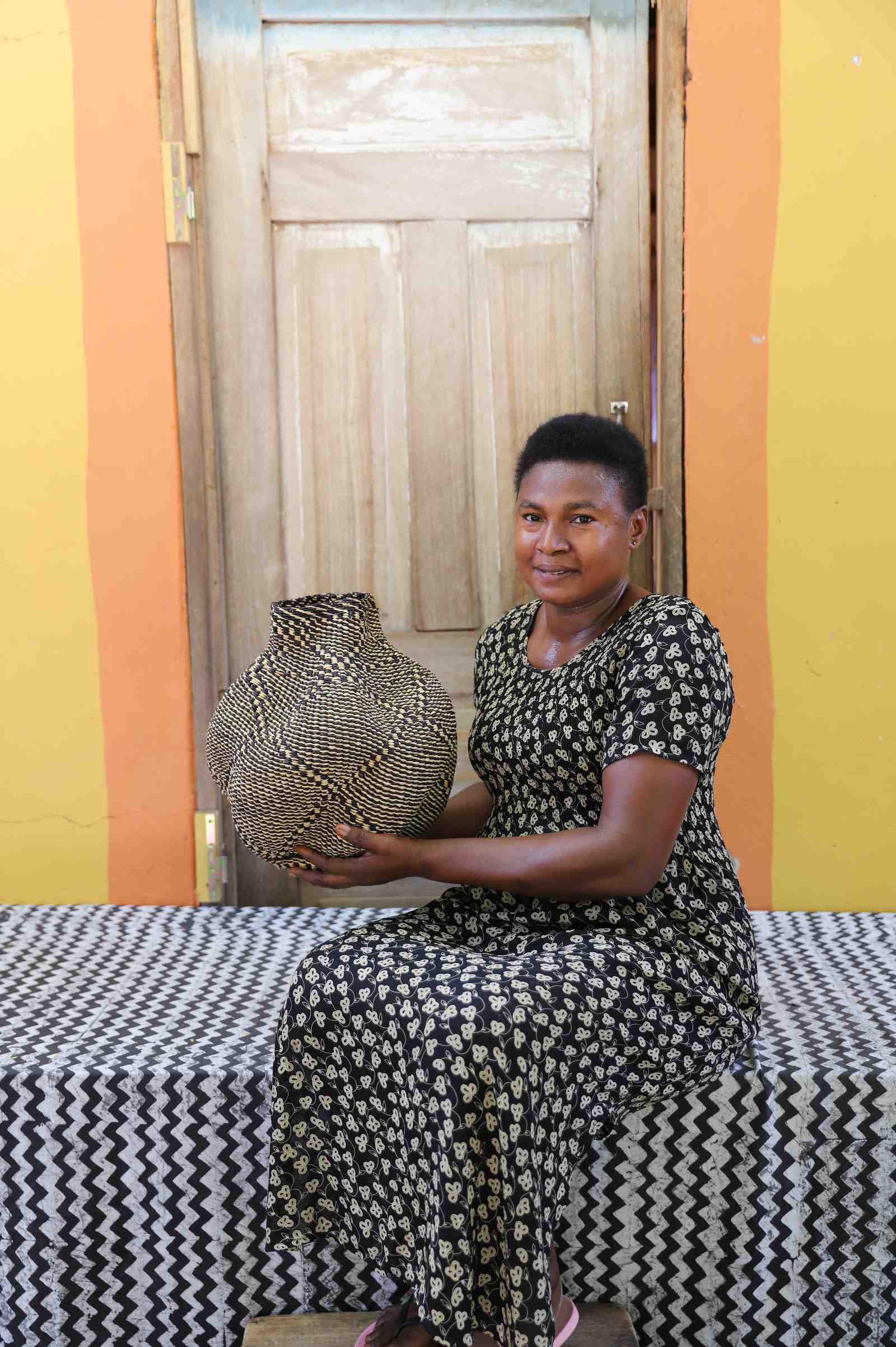 Handwoven Bolga art basket in natural straw with a fine dark diamond weave, held by a seated woman.
