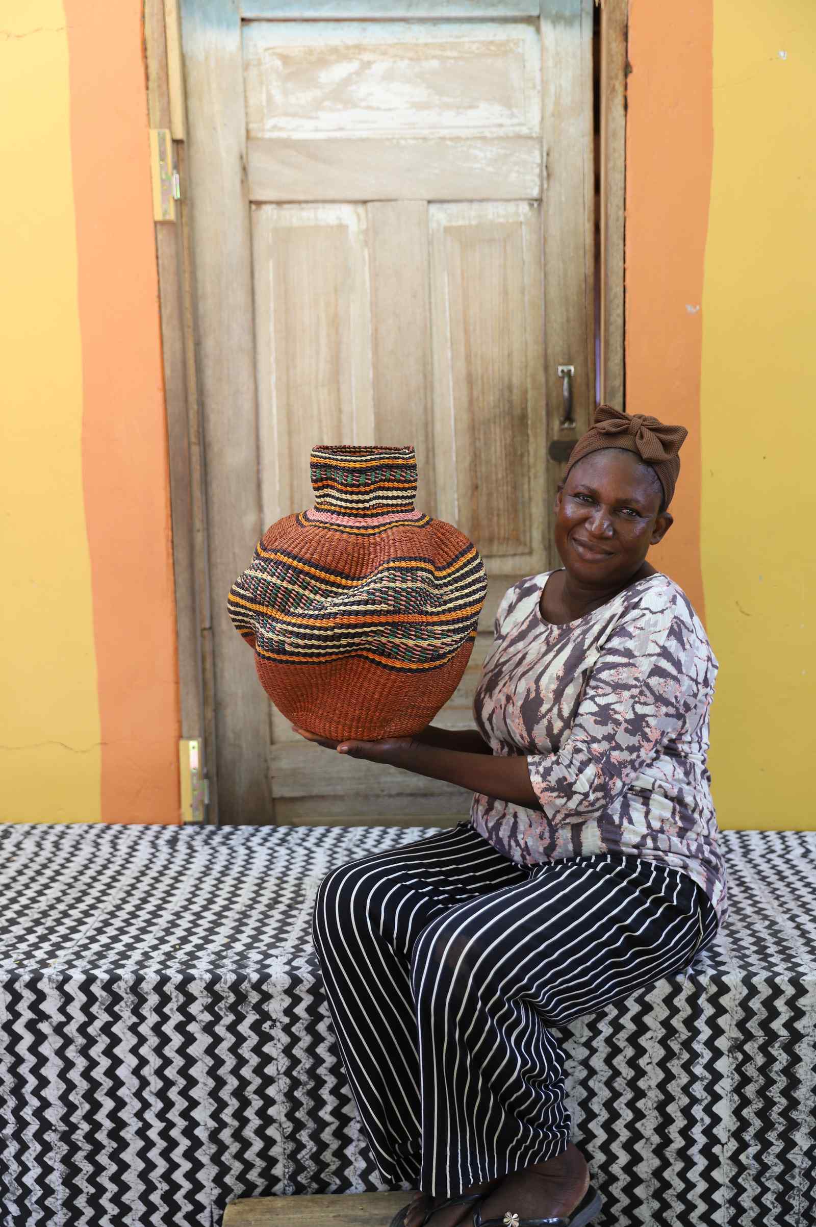 Handwoven Bolga art basket in rust-orange with a fine multicolour band, held by a seated woman in stripes.