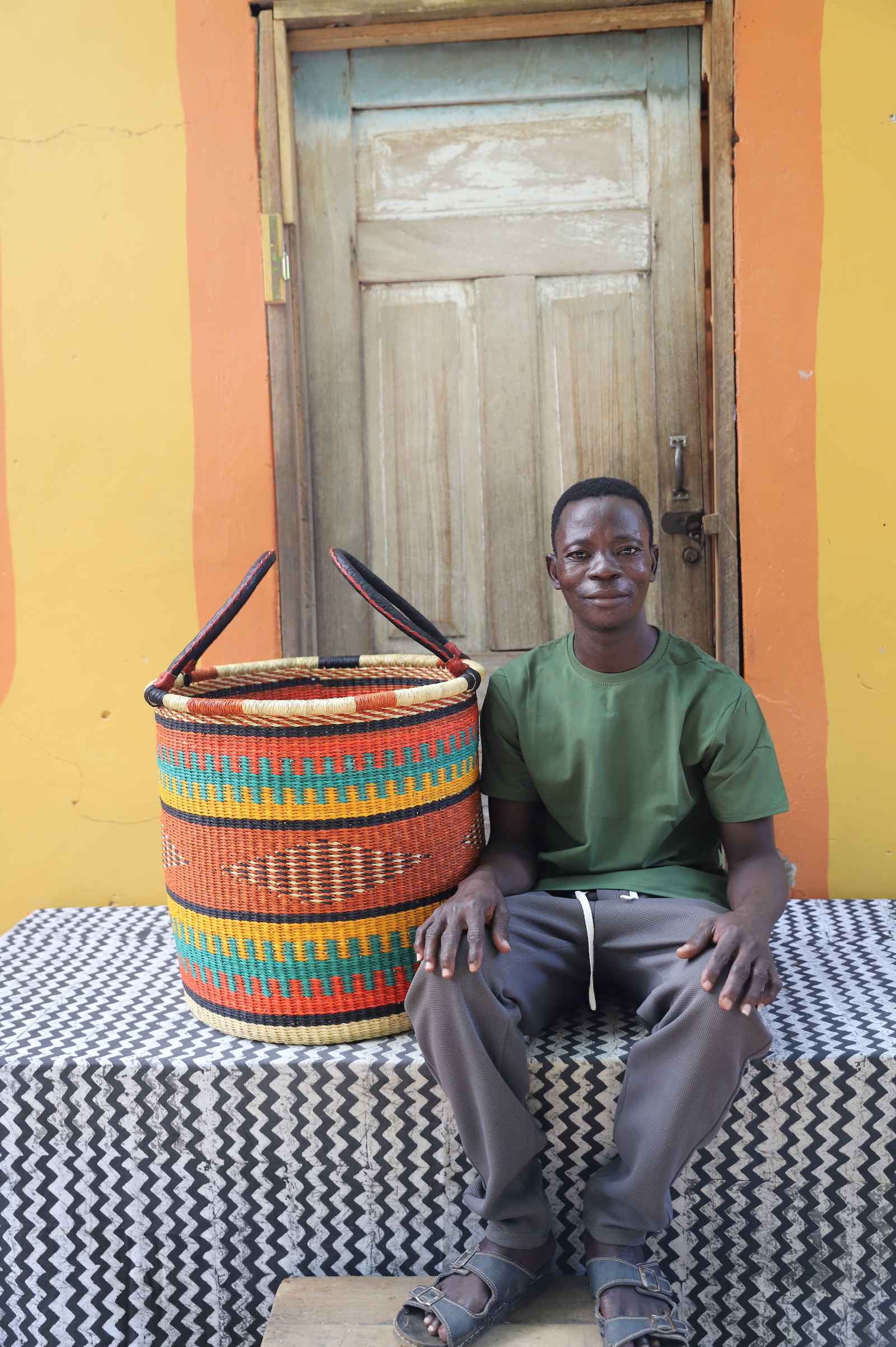 Large handwoven Bolga laundry basket in natural with orange and teal bands, beside a seated woman in floral.