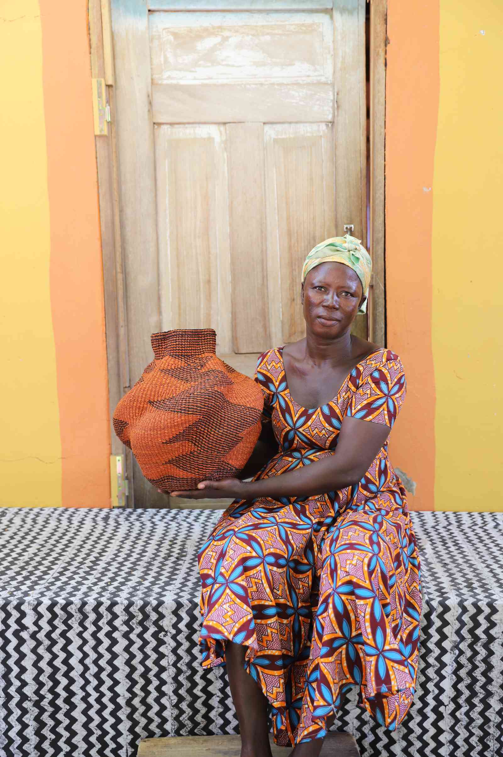 Handwoven Bolga art basket in rust-orange with a black arrow motif, held by a seated woman in a printed dress.