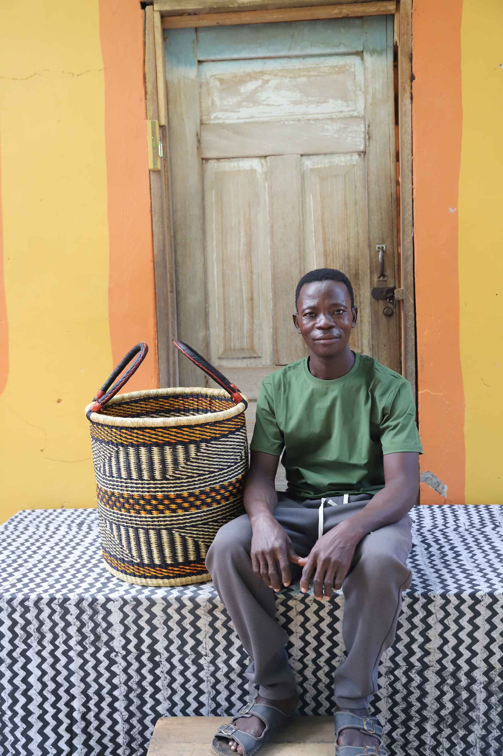 Small handwoven Bolga laundry basket in natural with fine dark stripes, beside a seated woman in a printed dress.