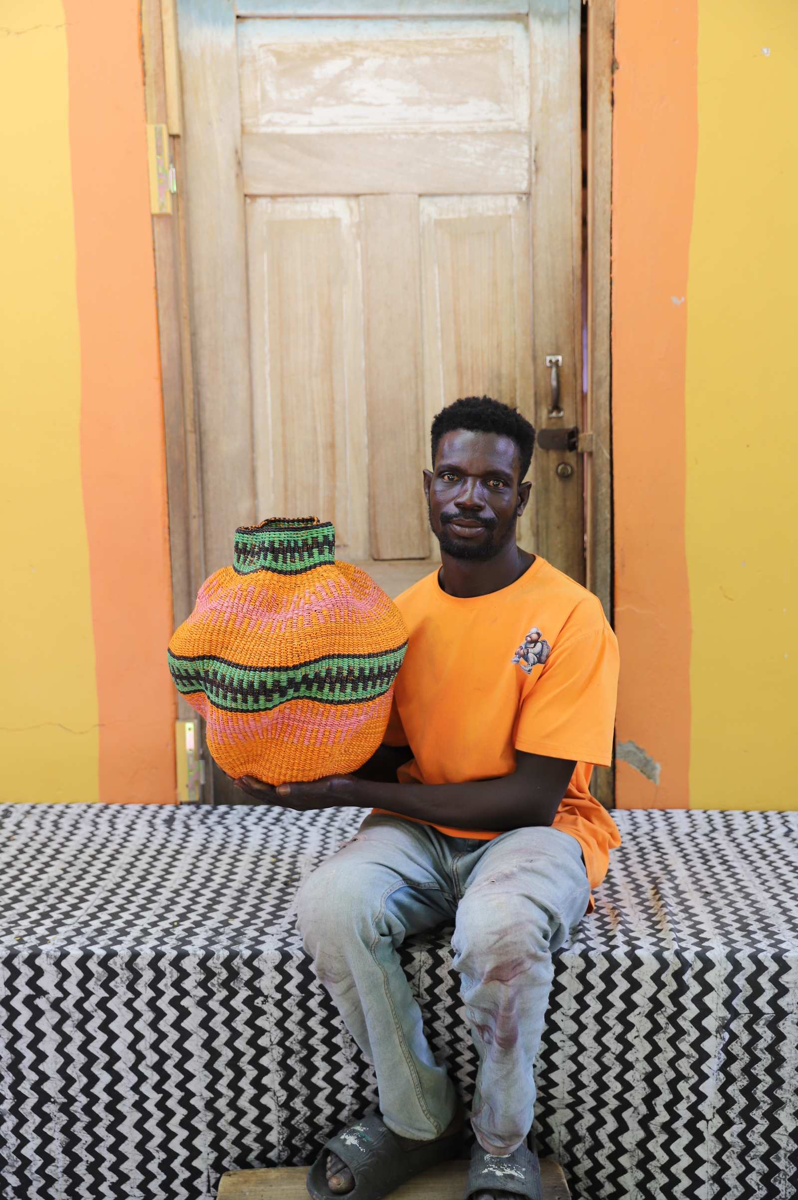 Handwoven Bolga art basket in dyed orange with green banded rim, held by a seated man in orange.