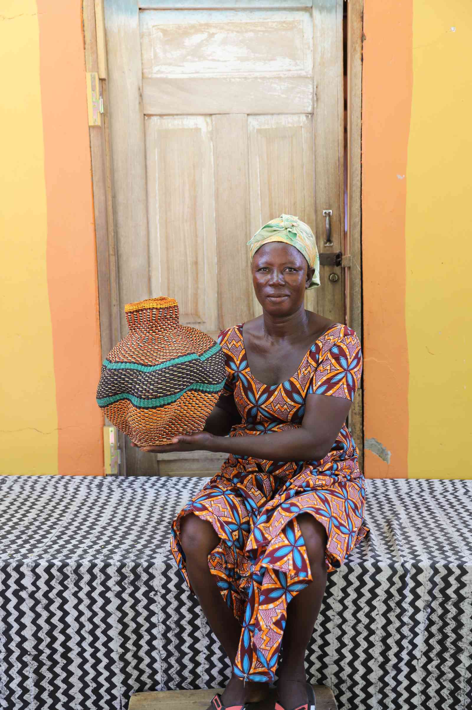 Handwoven Bolga art basket in natural straw with orange and teal bands, held by a seated woman in a patterned dress.