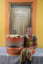 Handwoven Bolga laundry basket in rust-orange with a dark wave motif, styled beside a seated woman.