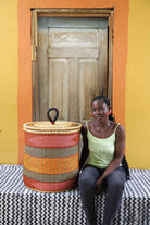Handwoven Bolga laundry basket in red, orange and yellow bands, styled beside a seated woman in green.