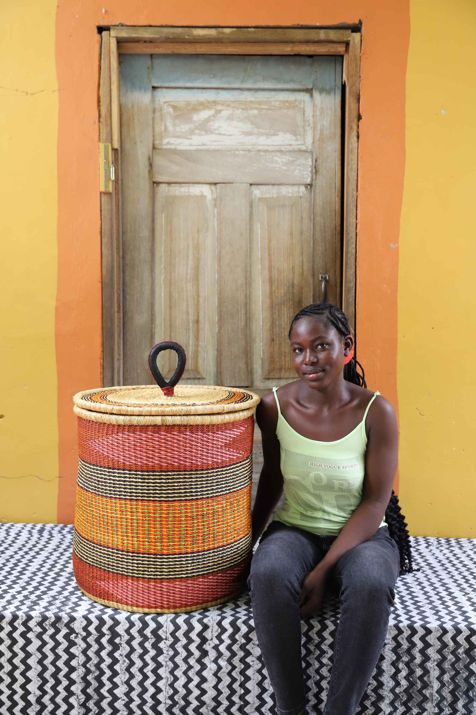 Handwoven Bolga laundry basket in red, orange and yellow bands, styled beside a seated woman in green.