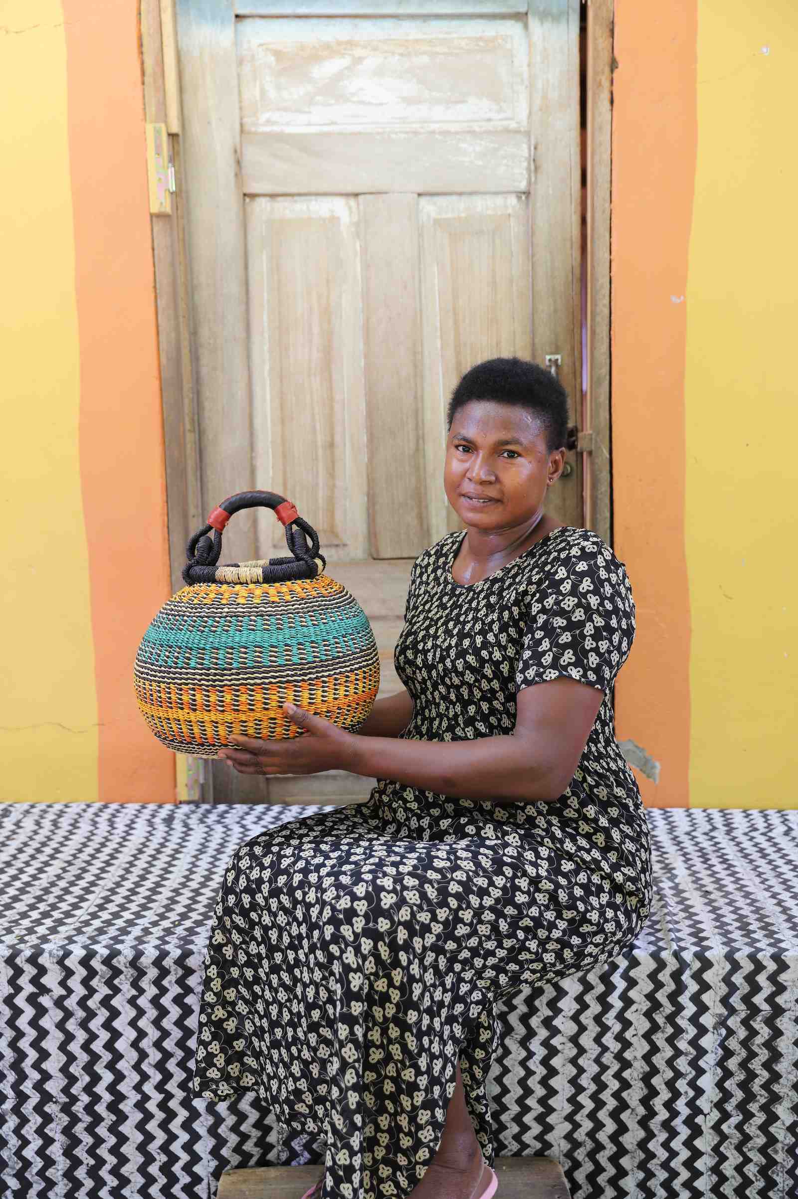 Handwoven Bolga pot basket with orange and teal bands, held by a seated woman in a patterned dress.