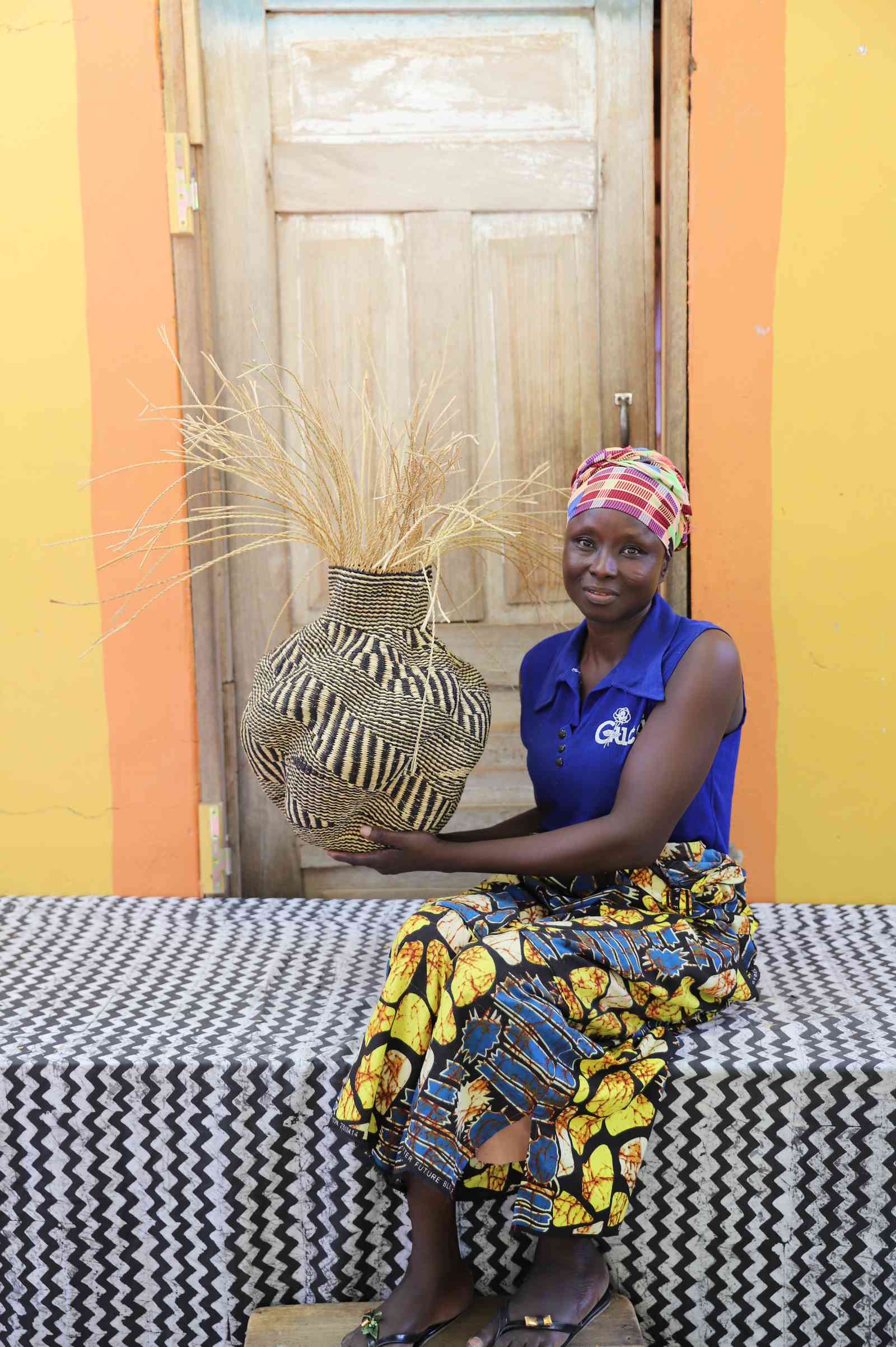 Handwoven Bolga Baobab basket in natural straw with dark trunk motif, held by a seated woman in a printed dress.